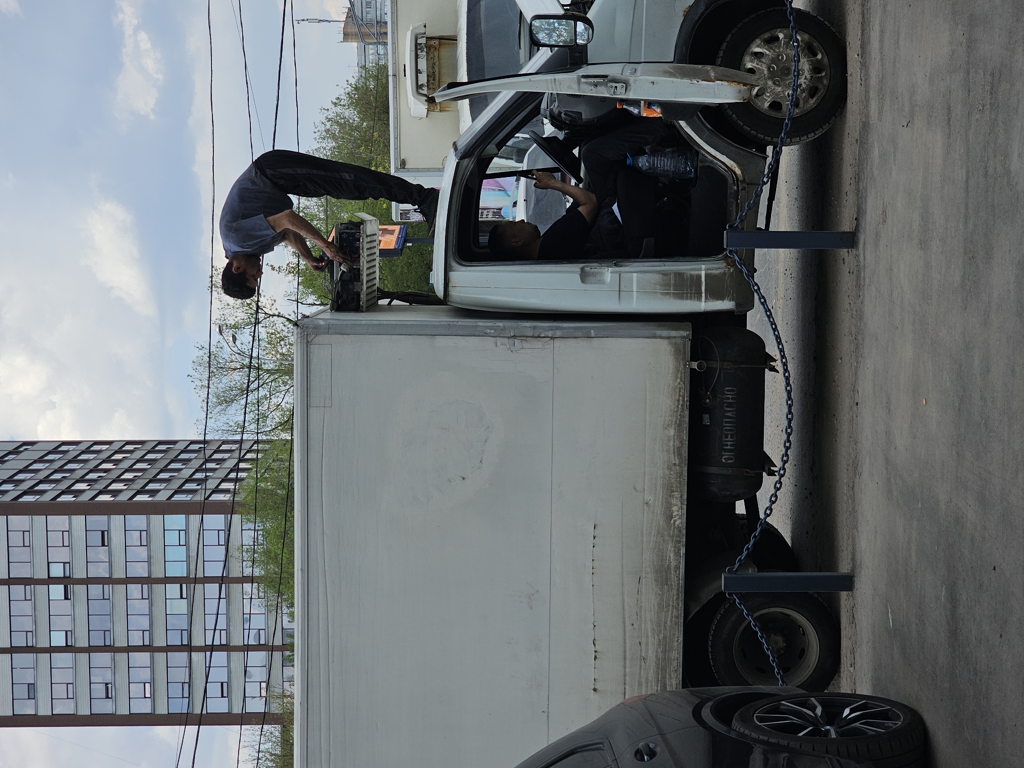 A medium-shot outdoor photo shows two men working on a vehicle in what appears to be Moscow, Russia. The image is oriented vertically.
The primary subject is a white commercial truck with a box-like cargo area. One man is positioned on top of the cargo box near the cab, bending over to work on an electronic device. He wears a gray shirt and dark pants. Another man sits inside the driver's seat, dressed in a black shirt.
The truck is parked on an asphalt surface, with the front of the vehicle angled towards the upper right corner of the image. A black car is partially visible in the bottom of the frame near the rear of the truck. A chain is seen securing the truck, running from the front tire to a steel beam or post.
In the background, there is a tall building with a grid-like pattern of windows, typical of urban architecture in Moscow. Overheard are some power lines. The sky is visible above the building, showing partly cloudy conditions. Green trees are visible in some background.