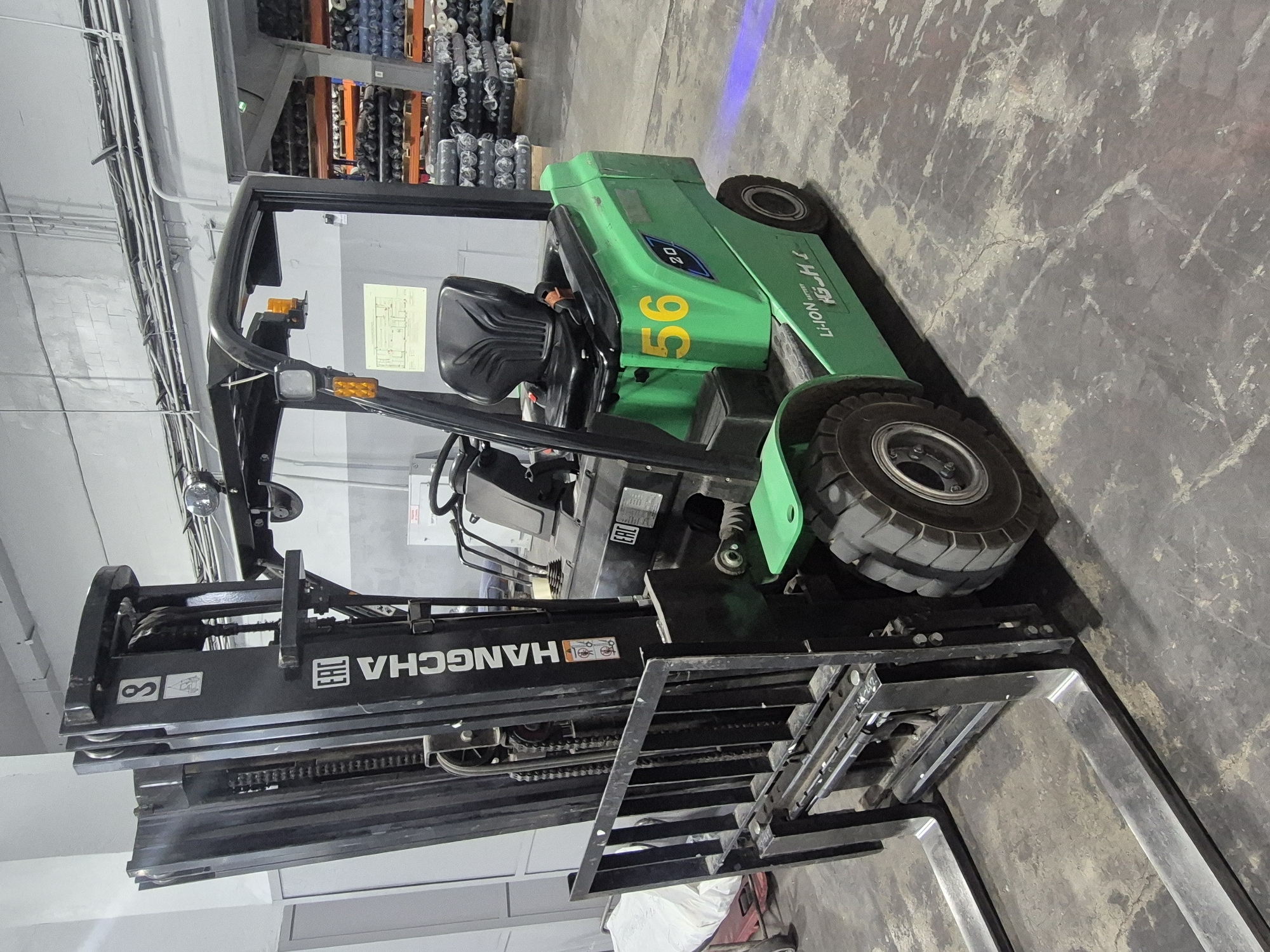 A green Hangcha forklift with the number 56 on the side sits in a warehouse. The word "HANGCHA" is visible on the front of the forklift. Racks of materials are in the background. The location is in a warehouse in Chaikovsky, Russia.