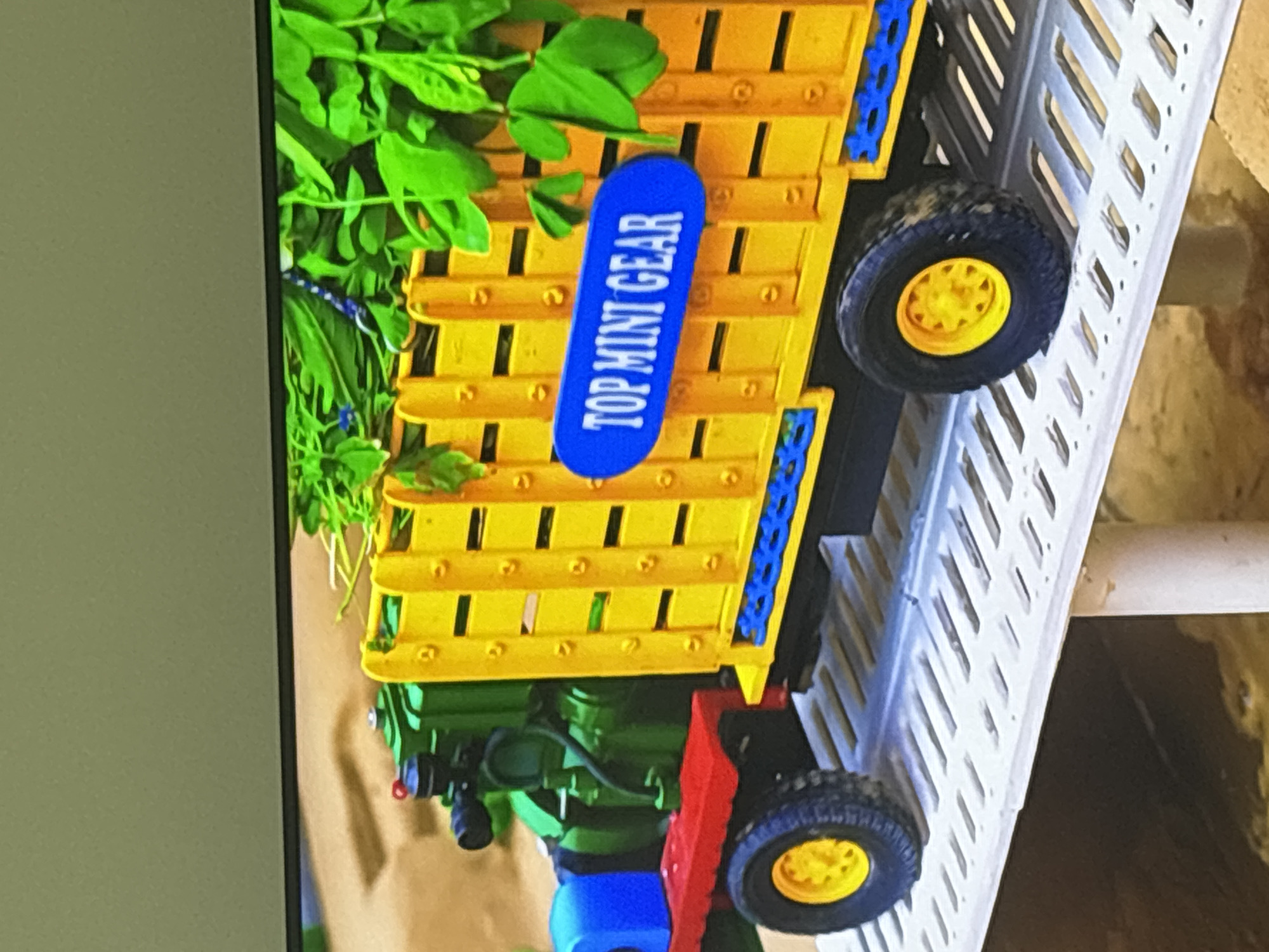 The image shows a close-up, side-view of a toy truck. The truck is green and red in the front, with a blue compartment. The back of the truck is yellow and designed like a trailer with vertical slats and a blue decorative trim at the bottom. A blue oval sign with white text that reads "TOP MINI GEAR" is attached to the side of the yellow trailer. Green leafy plants are visible in the back of the trailer. The truck is positioned on a white, slatted platform. The background appears to be a blurred outdoor environment with sandy or dirt-like ground. There are no people visible in the image. The image does not contain any information that allows for its location to be determined as Dubai, United Arab Emirates.