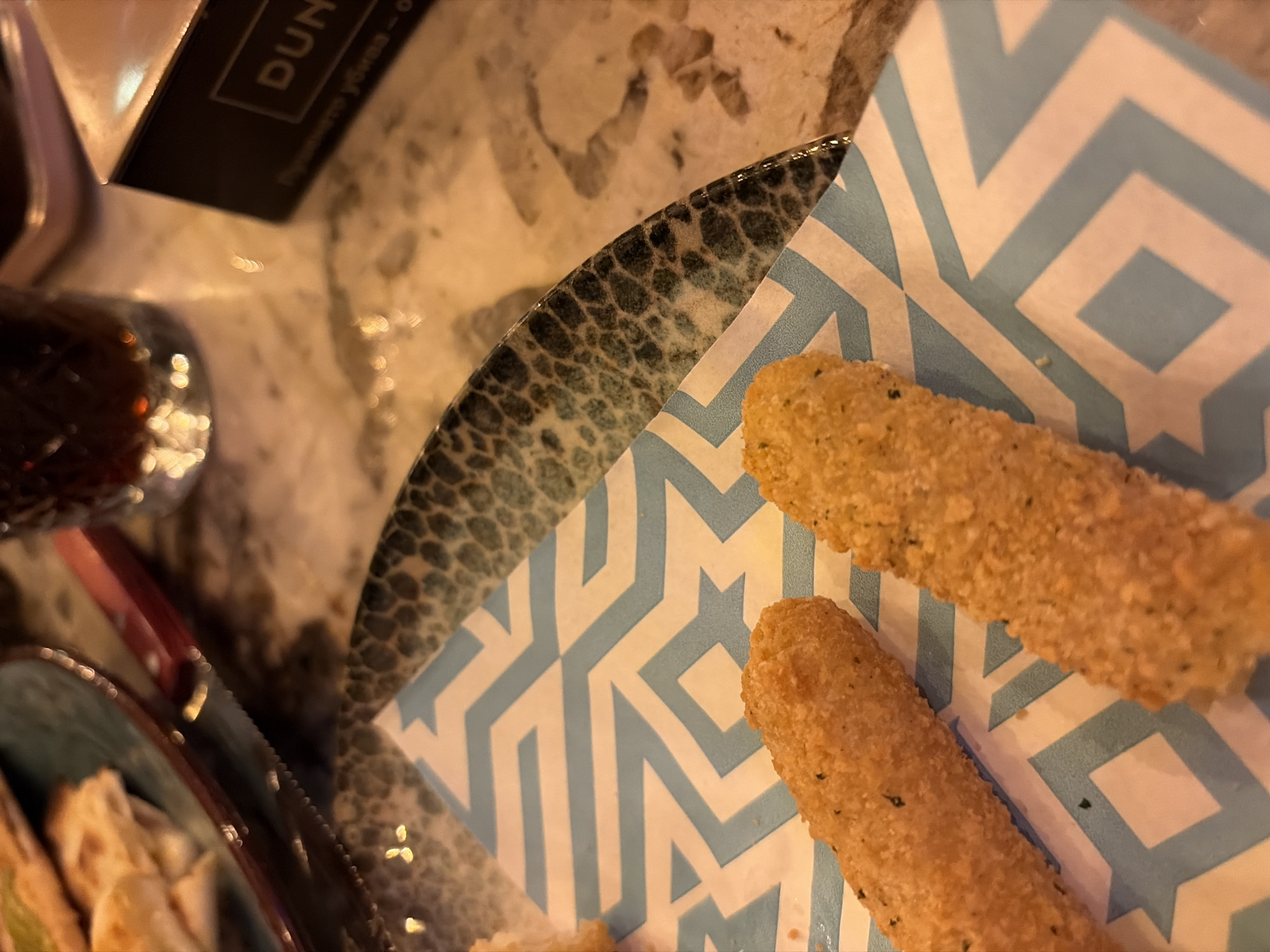 The image shows a plate of fried mozzarella sticks on a geometrically patterned blue and white paper napkin, placed on a mottled grey surface, potentially a table. A knife and fork are visible on the lower left, along with part of another plate containing some food. In the background, there's a glass with a dark liquid and a container, possibly a menu holder, with the word "DUN."
Given the presence of Cyrillic text ("пушено убива") near the word "DUN," it is likely that the location is in Bulgaria. Considering Bulgaria's capital city is Sofia, the scene may be located within the city of Sofia. Without further information, I cannot confirm the specific restaurant or establishment. The focus is on the food item, suggesting a dining or eating environment.