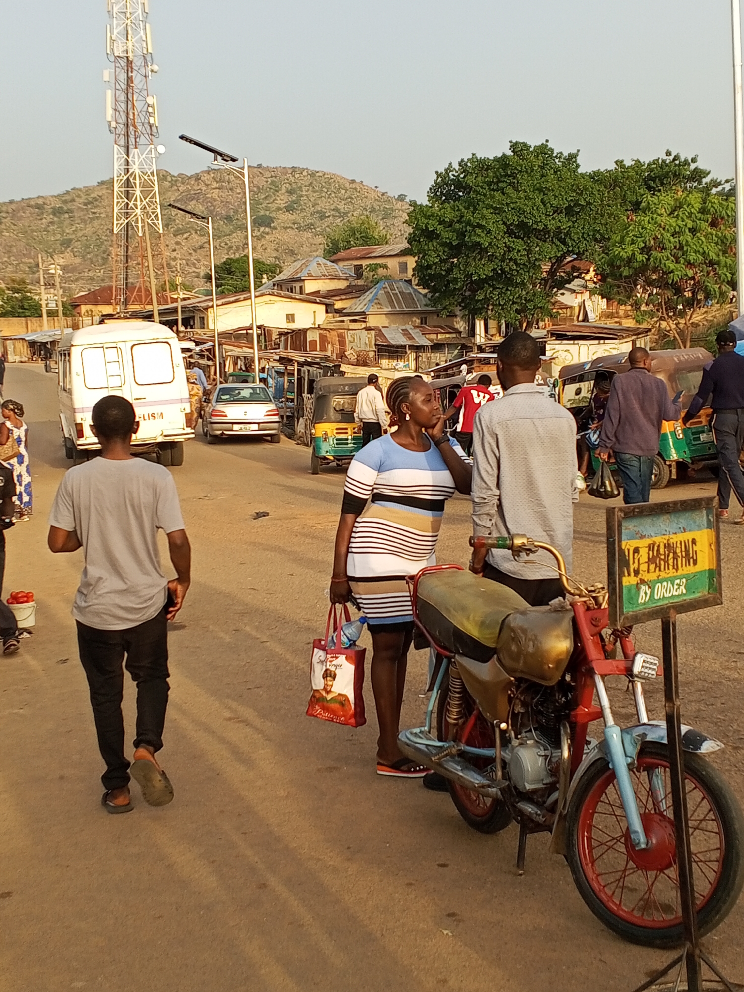 The image depicts a street scene in Gwong, Nigeria. The location is characterized by a paved road and various buildings and structures, including a cell tower and a hill in the background. Several people are present, including a woman in a striped dress who appears to be talking to a man standing next to her. A motorcycle is parked nearby, next to a sign that reads "No Parking by Order." A man in a gray t-shirt and black pants is walking away from the scene. Other vehicles, including a white van, a car, and several auto-rickshaws, are visible in the background. Additional people are present in the background as well.