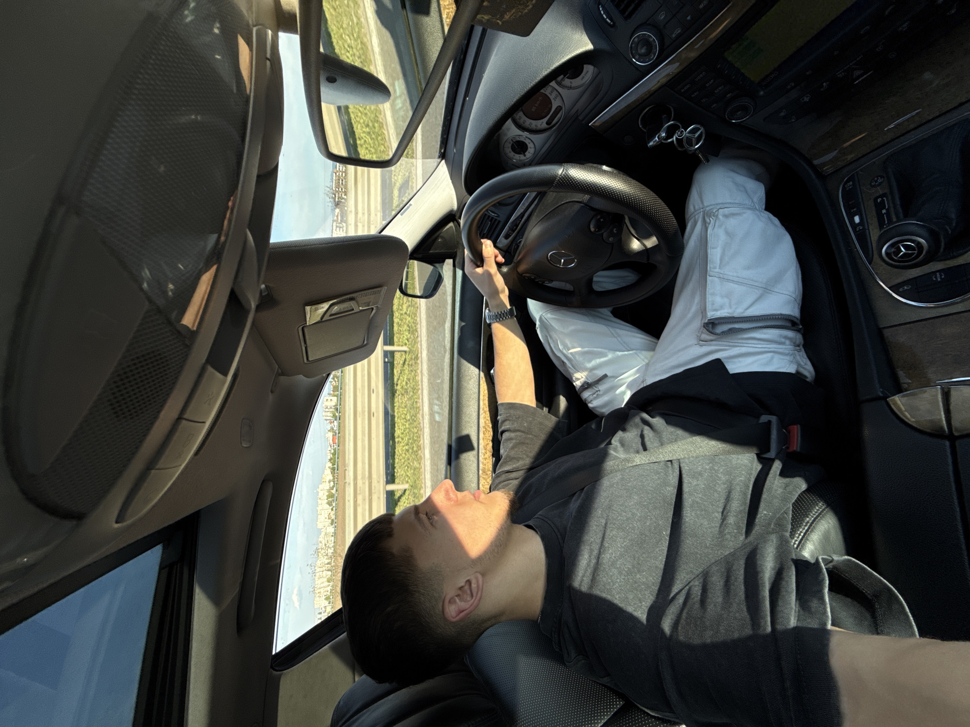 A young man is reclining in the driver's seat of a Mercedes-Benz vehicle. He wears a gray t-shirt and white pants, with his feet up on the dashboard. The car is located on a road or highway in Sofia, Bulgaria. The scenery outside the windows reveals buildings and infrastructure typical of an urban environment.
