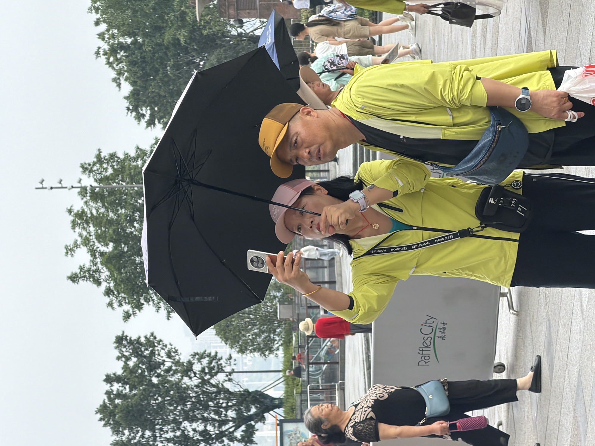 In a city environment, likely Yuzhong District, China, on a cloudy day with overcast skies, two individuals are captured under a large black umbrella, shielding themselves from the weather. The primary focus is on a woman in the foreground, wearing a bright yellow hooded jacket and a pink baseball cap. She is holding a smartphone in her right hand, taking a selfie. She also wears a turquoise top underneath her jacket and has a red pendant necklace. On her left wrist, a smartwatch is visible. Next to her, a man is looking off to the side, wearing a similar bright yellow hooded jacket over a black t-shirt and a tan baseball cap. He has a blue crossbody bag slung over his shoulder and is holding a white plastic bag in his right hand. Both individuals appear to be walking or standing in a paved outdoor area, possibly a public square or promenade, with trees and buildings visible in the background. A large gray barrier with "Raffles City" branding is present to the left of the frame, indicating a commercial or entertainment district. Other people, including an older woman in a black top and black pants with a blue shoulder bag, are visible in the mid-ground and background, some carrying umbrellas, suggesting it might be raining lightly or that rain is expected. The overall scene depicts everyday urban life, with people navigating the environment and using technology.