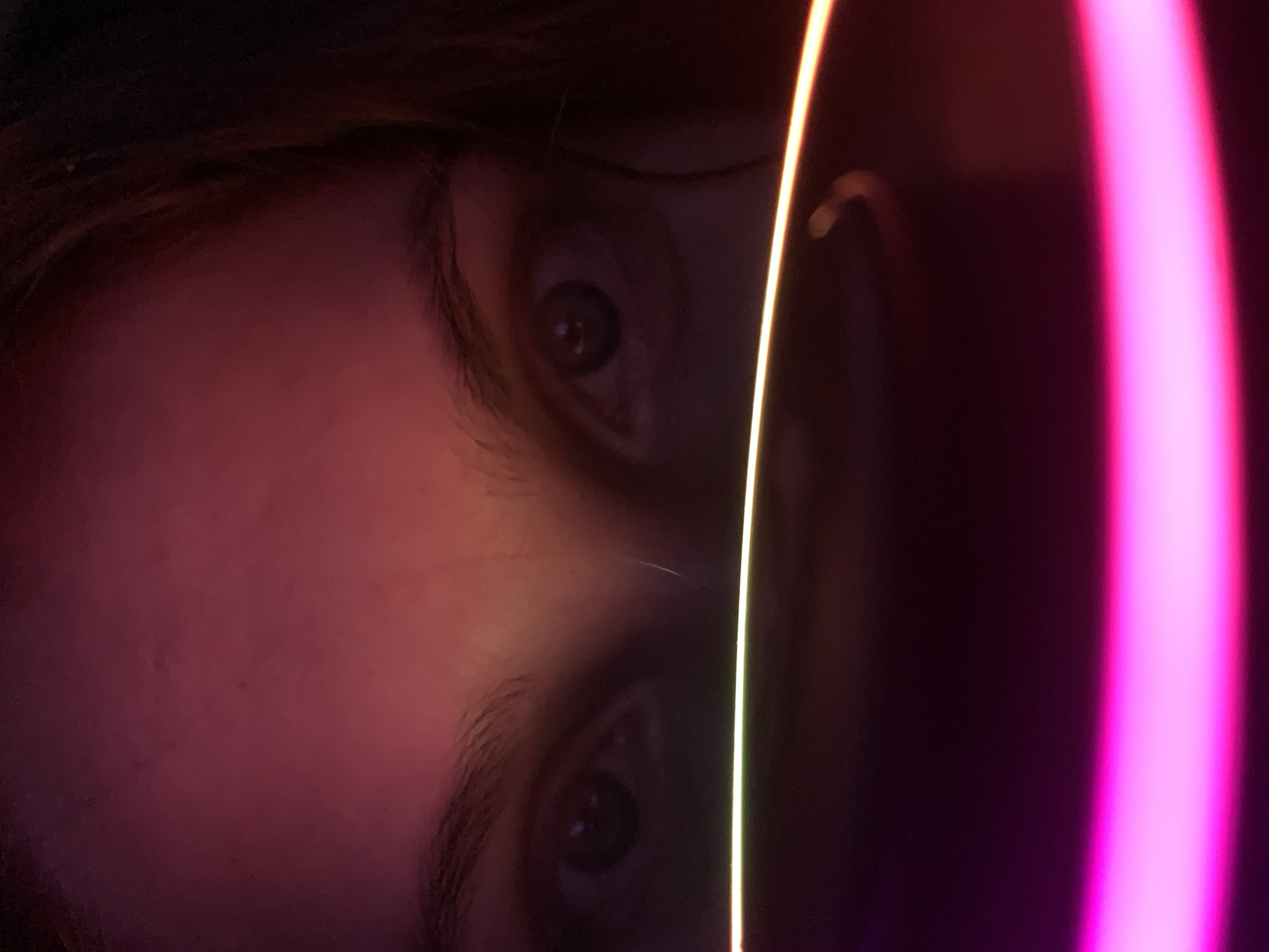 The image displays a close-up, low-light portrait of the upper portion of a person's face. The subject's eyes, forehead, and parts of the hair are visible, illuminated by strong, artificial colored lights. The person's eyes, appearing dark and with somewhat dilated pupils, are directed forward. Dark eyebrows are visible above the eyes. Dark hair frames the face on the left and right sides. The facial skin tone appears light to medium, but it is heavily tinted by the ambient lighting, which casts a predominant reddish-pink hue across the forehead and eye areas.
Prominently, two distinct light trails horizontally bisect the lower half of the image. The upper trail is a thin, bright line, primarily yellow-green, positioned just below the subject's eyes and across the perceived bridge of the nose. Below this, a wider, intensely bright magenta/pink light trail curves across the bottom of the frame. A fainter, curved orange light streak is also visible in the mid-right portion of the image, above the magenta trail. These light trails suggest motion blur from a moving light source or a long exposure photograph. The background is indistinct and dark, with subtle gradients of red and orange indicating the broader influence of the colored light sources. The specific location in the city of Chaikovsky, Russia, cannot be determined from the visual information present in the image.