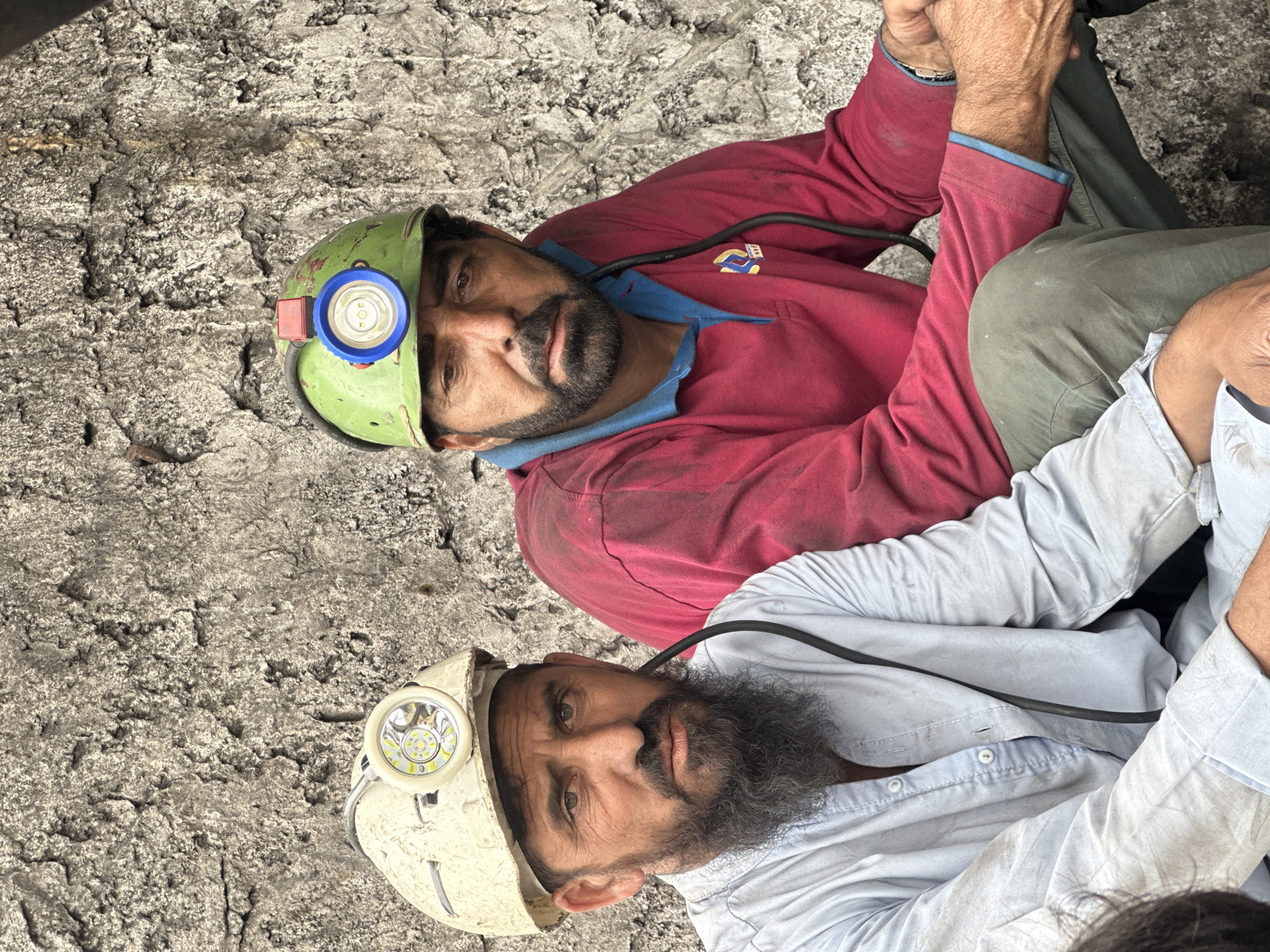 Two men, likely coal miners, are seated in front of a rough, grey, textured wall, possibly in a mine setting in Quetta, Pakistan. The man on the left wears a light blue, button-down shirt and a white hard hat with a headlamp. He has a full, dark beard and appears to be looking directly at the camera with a serious expression. A black cable runs from his hard hat. The man on the right wears a red long-sleeved shirt with a blue collar and a green hard hat with a headlamp. He has a beard and appears to be looking downwards, with a tired or contemplative expression. He is wearing green pants. Both men's clothing appears worn and possibly dusty, consistent with manual labor. The wall behind them is uneven and shows signs of weathering. The lighting suggests an outdoor or dimly lit environment, with the headlamps providing localized illumination.