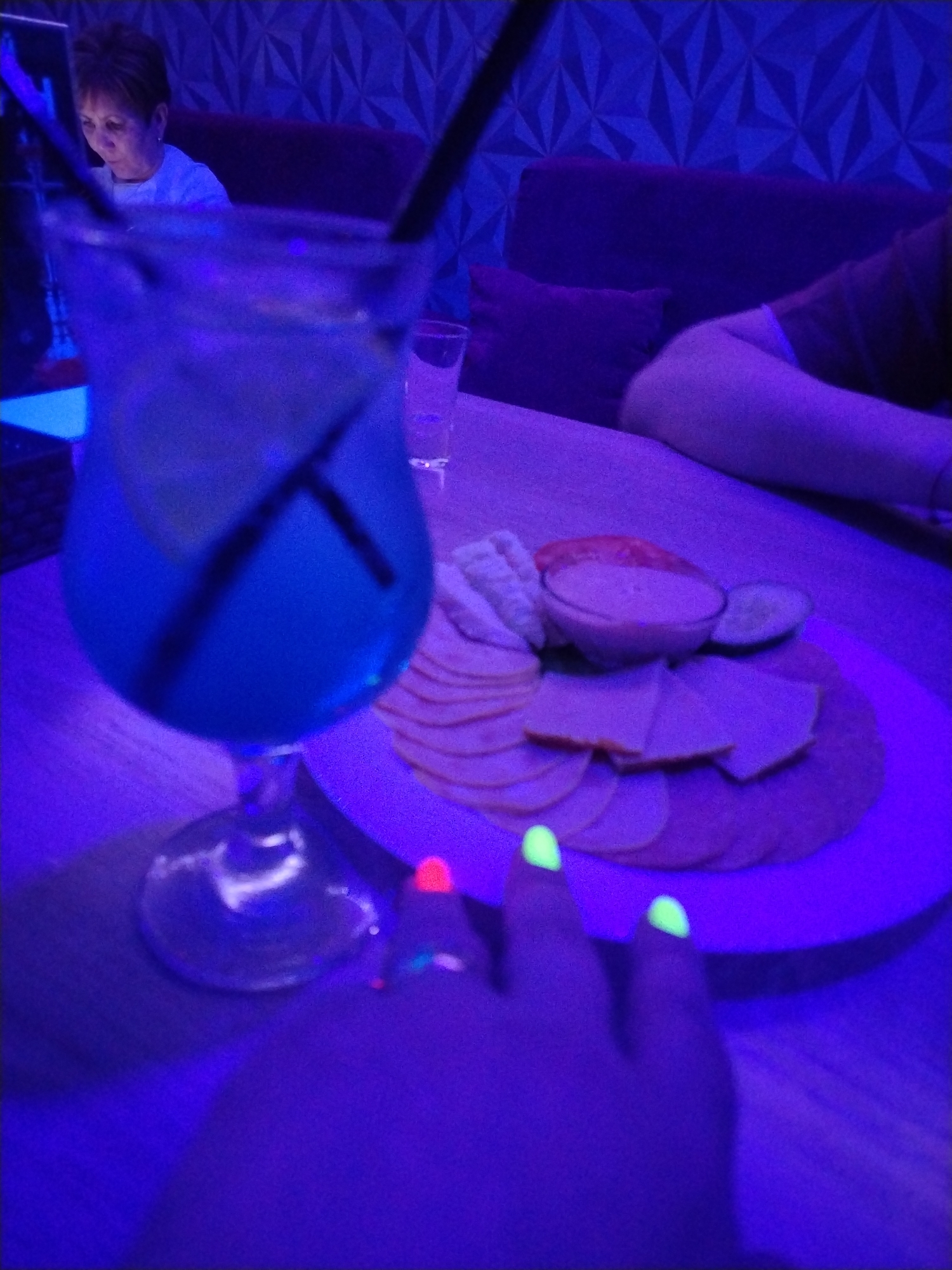 The image captures an indoor evening scene in Chaikovsky, Russia, likely a lounge or bar, characterized by strong blue and purple ambient lighting, possibly from UV or blacklight, which gives a glowing effect to certain objects.
In the foreground, a tall, stemmed glass holds a vibrant blue beverage with two dark straws and a citrus slice, appearing to glow under the lighting. To its right, a clear glass contains water. A person's hand, presumably the photographer's, is prominent in the bottom right, with fingernails painted in bright, glowing neon green and pink, indicating the presence of UV light.
A round plate of snacks is centrally placed on the table, featuring various sliced meats or cheeses, crackers, a small bowl of dip, and cucumber slices. The food items appear muted in color due to the predominant blue light.
In the mid-ground and background, a woman with short brown hair, wearing a light-colored top, is visible, looking down. Her face is partially obscured by the blue drink. She appears to be sitting on a dark sofa or bench. To her right, part of another person's arm and shoulder can be seen resting on the same furniture. The background wall features a distinctive textured, geometric pattern.
The overall atmosphere suggests a relaxed, social gathering, with people enjoying drinks and snacks in a uniquely lit environment. No discernible text is visible in the image.