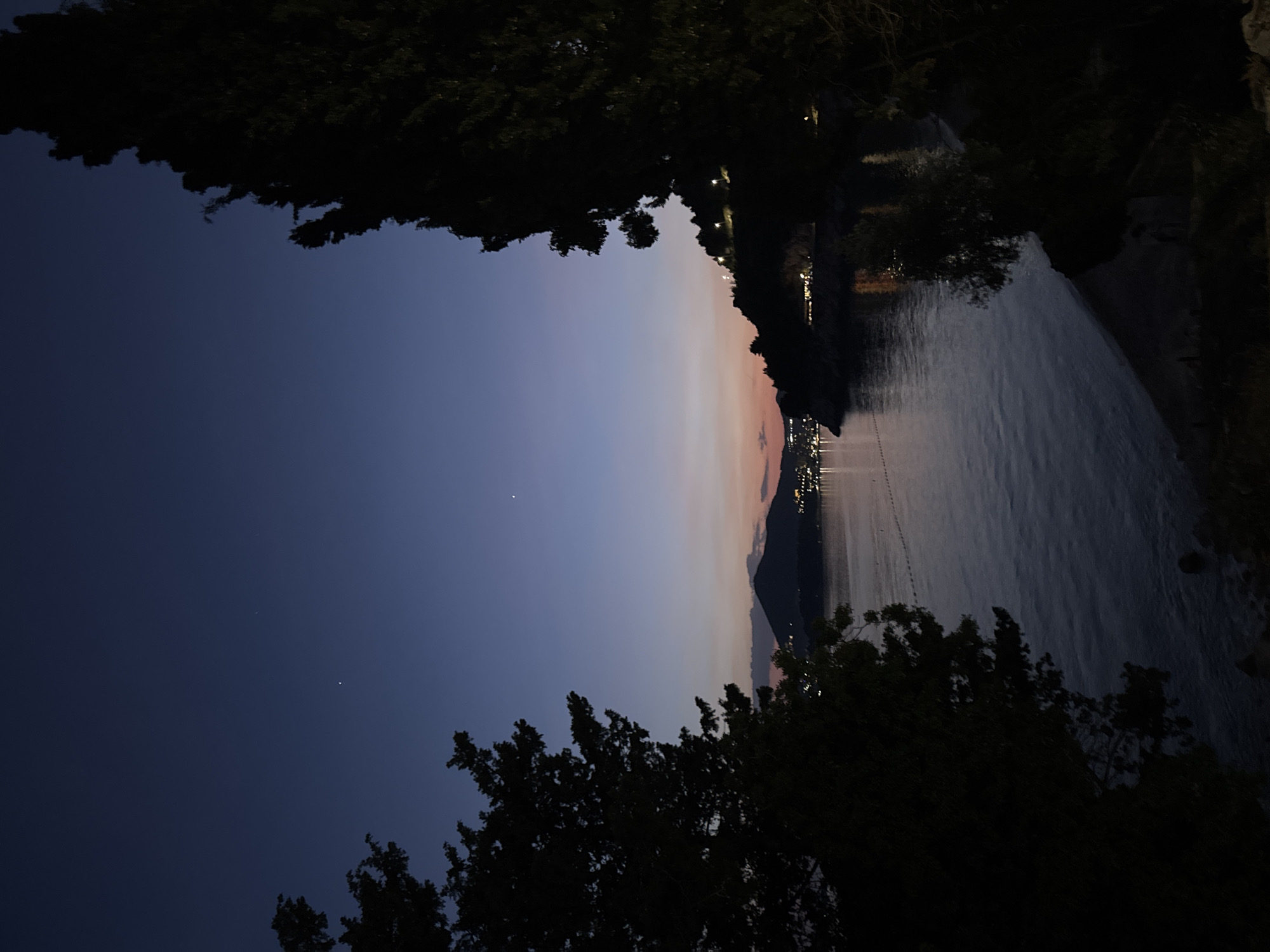 The image shows a nighttime view in Kamenovo, Montenegro. The sky is a gradient from dark blue to a pale pinkish hue near the horizon, where faint city lights are visible. A body of water, possibly the Adriatic Sea, fills the right side of the frame. The edges of the frame are obscured by dark tree silhouettes. The scene is dark, with limited details visible due to the low light. There is no evidence of people or animals.