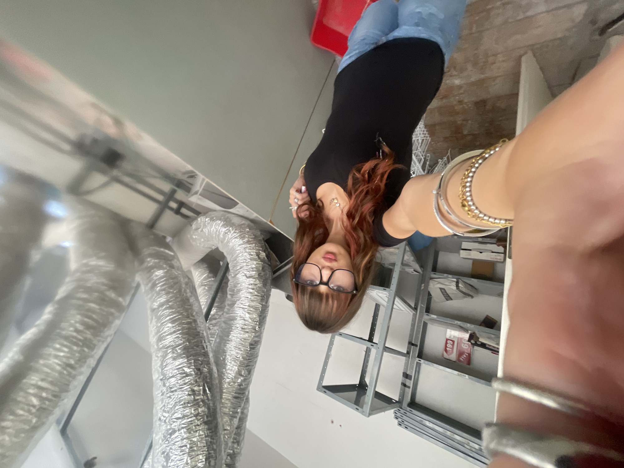 A medium shot captures a young woman with shoulder-length brown hair, styled with bangs and reddish highlights, wearing black-rimmed glasses and a black scoop-neck t-shirt, taking a selfie. She is positioned in what appears to be a storage or workshop area, with shelves filled with various items in the background, including what looks like inventory boxes and signage. Large, silver, flexible ventilation ducts dominate the right side of the frame. The floor is made of wooden planks. The woman is adorned with several bracelets on her left wrist and rings on her fingers. Her right arm is extended, holding the camera, with some of her hand visible in the foreground. The overall lighting suggests an indoor environment, possibly a commercial or industrial space. The context of Raanana, Israel, is provided, but there are no specific visual cues within the image to confirm this location.