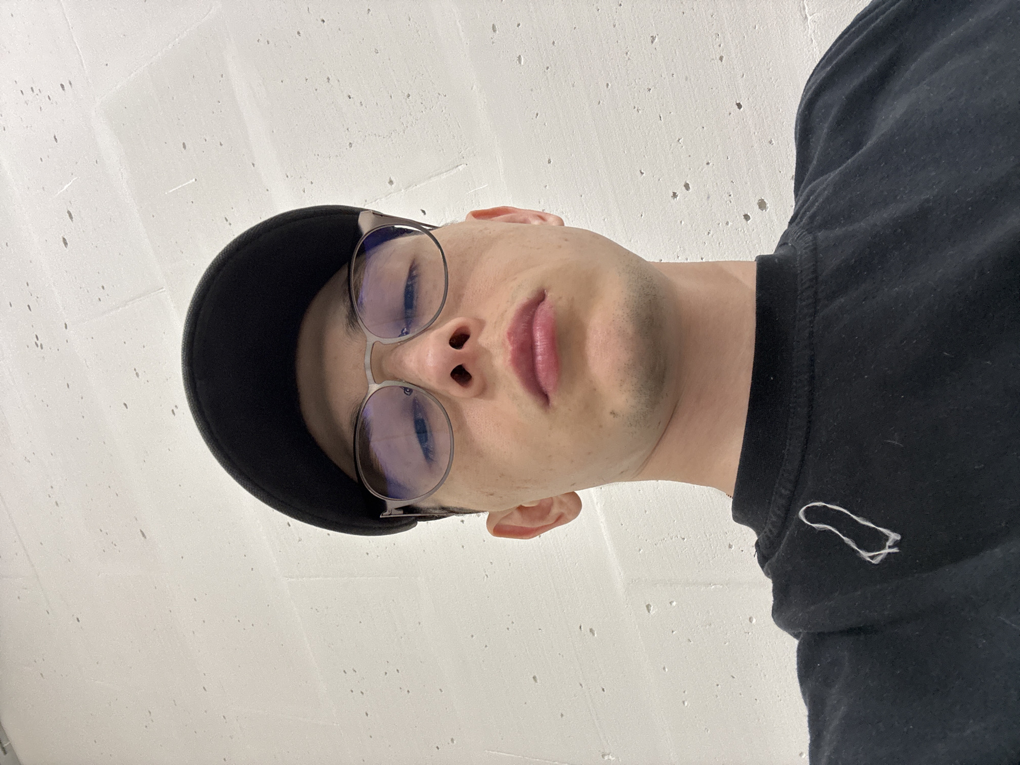 The image is a close-up, low-angle selfie of a young man wearing a black baseball cap and black t-shirt. He has on thin-rimmed glasses and his expression is neutral, with his lips slightly parted. A white, irregularly shaped object, possibly a piece of string or thread, is visible on the left side of his t-shirt. The background is a textured white wall. The scene appears to be indoors, possibly in a city like Ostrov, Russia, given the texture of the wall which resembles concrete common in urban environments. There is no indication of specific outdoor features or landmarks associated with Ostrov.