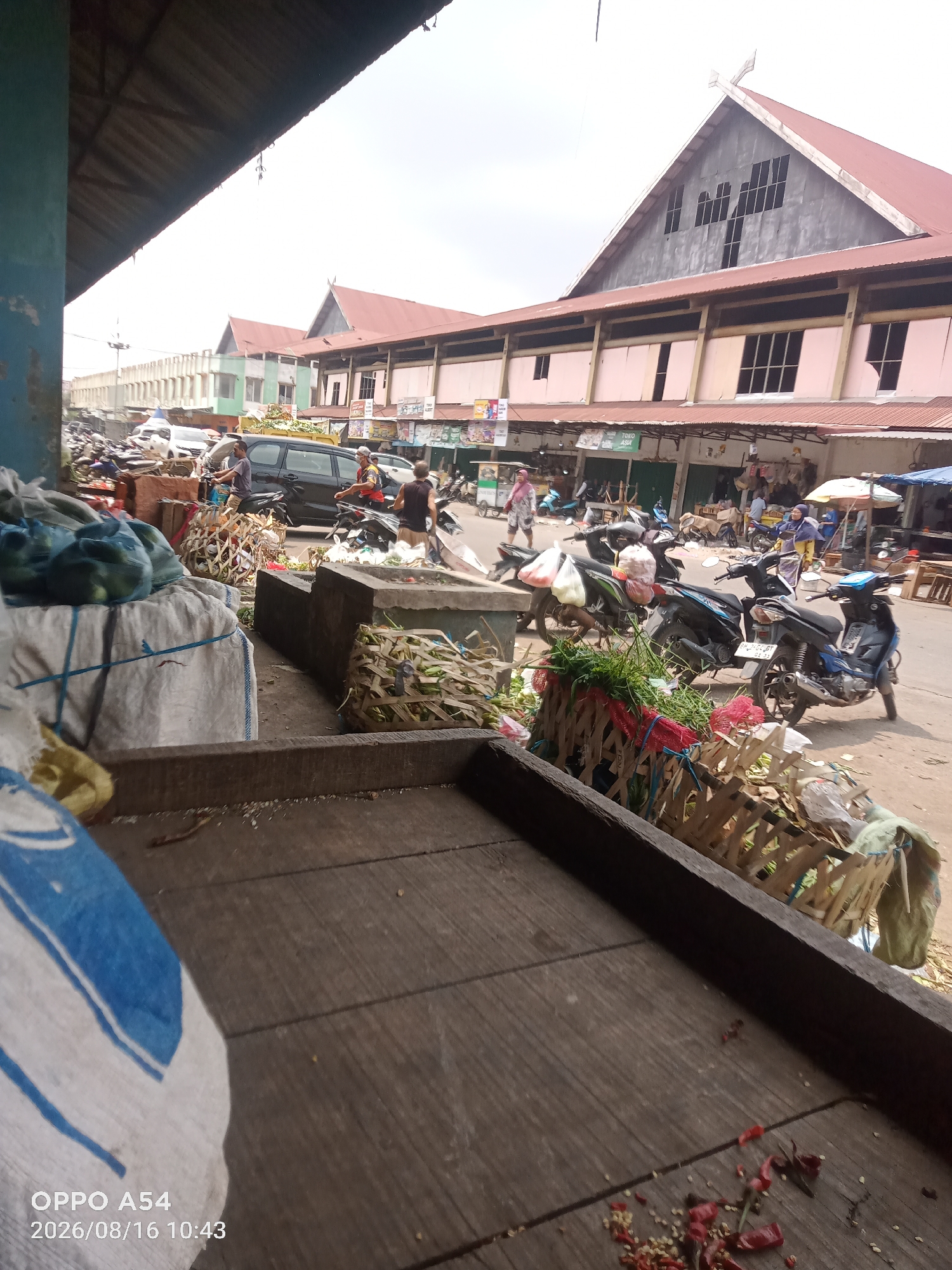 The image depicts a bustling market scene in the city of Jambi, Indonesia. The foreground is dominated by a wooden structure, possibly a market stall, with sacks of produce and scattered red chili peppers on its surface. Beyond this, the scene opens up to a street filled with various motorcycles and a black van. Several individuals are visible, some riding motorcycles and others walking through the market. In the background, a long building with a reddish-brown roof and numerous windows suggests the presence of shops or market stalls. The overall atmosphere is that of a busy commercial area.