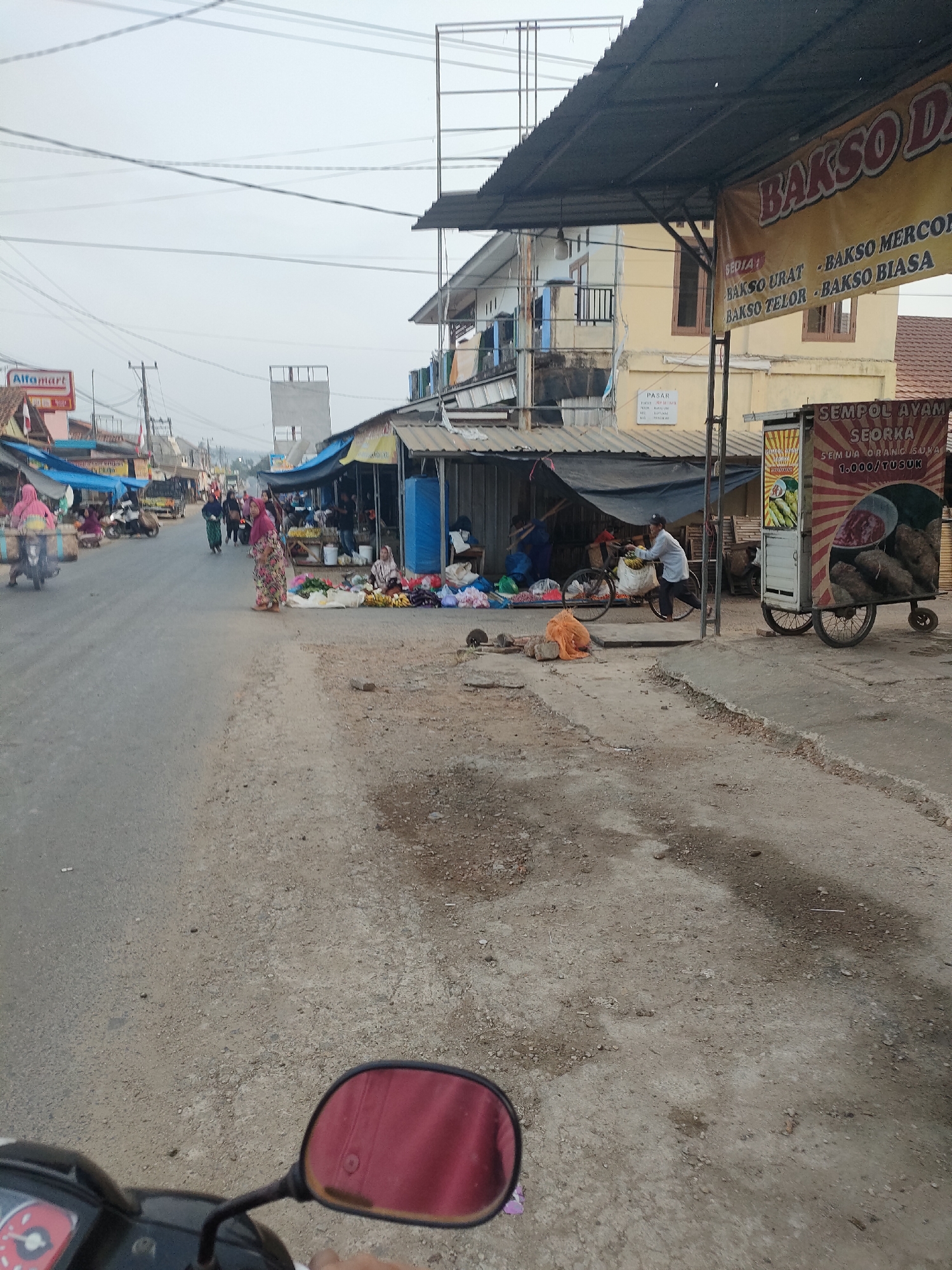 The image depicts a street scene in Sukoyoso, Indonesia, likely a market area given the presence of stalls and vendors. In the foreground, a portion of a scooter's handlebar and mirror is visible, suggesting the photo was taken from a moving vehicle. The road is paved with asphalt and appears somewhat worn, with patches of dirt and debris.
Several market stalls line the right side of the road, covered by dark awnings. One stall prominently displays a sign for "SEMPOL AYAM SEORKA" and advertises "1.000/TUSUK." This stall has a wheeled cart with a graphic showcasing fried chicken skewers and a dipping sauce. Another stall to the left has various produce, including bananas and vegetables, laid out on the ground.
People are scattered throughout the scene. A woman in a floral dress stands in the middle of the road, facing away from the camera. To her left, a man is pushing a bicycle laden with goods. Further down the street, several individuals are seen walking or standing near the market stalls. On the left side of the road, a few more stalls are visible, and a woman on a scooter is passing by. The background shows buildings, including a multi-story structure with yellow walls and balconies, and a sign for "ALFAMART" further down the street. The sky is overcast, suggesting a cloudy or hazy day. The overall atmosphere appears to be a bustling local market.