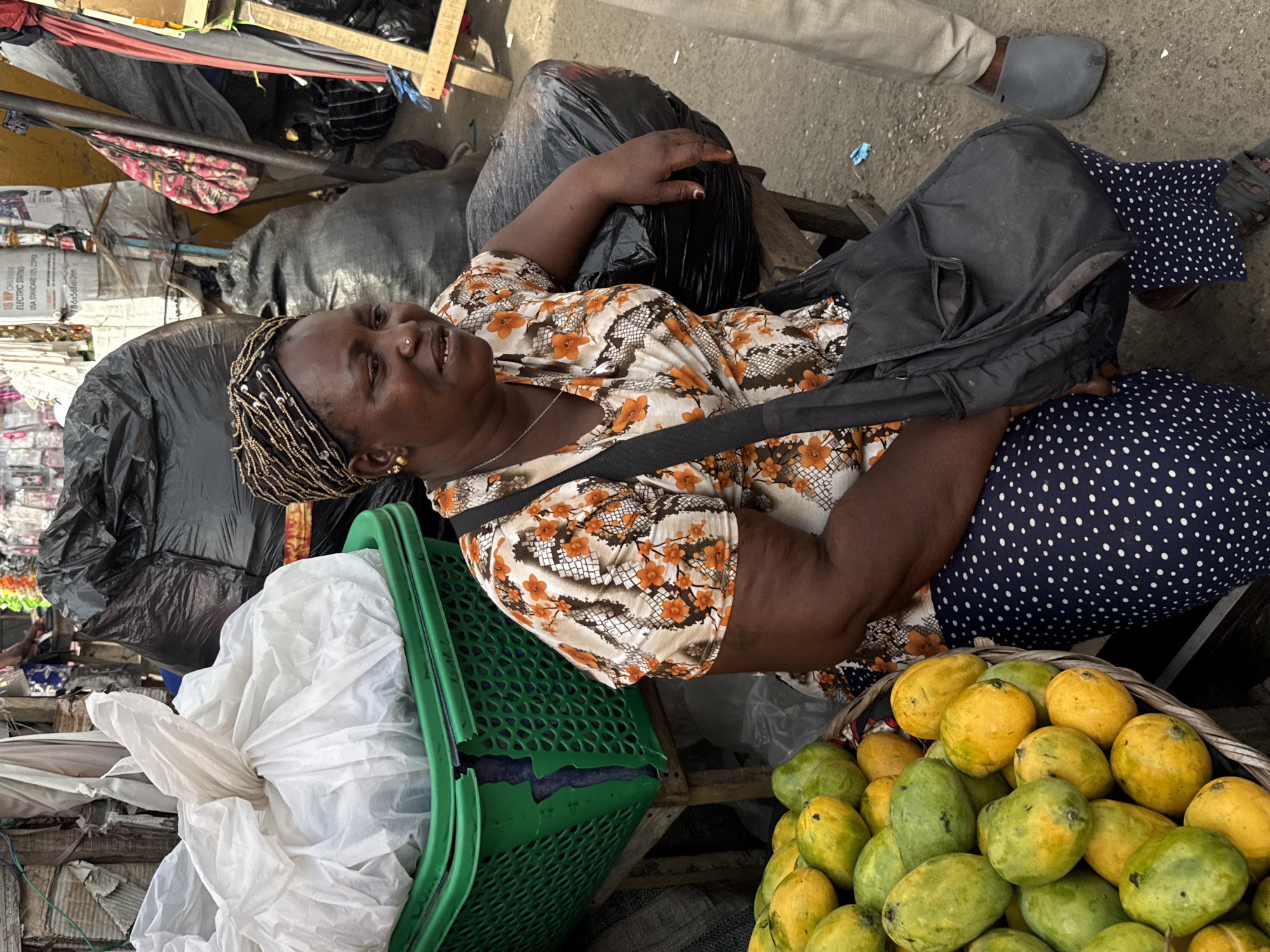 The image captures a seated woman within a bustling, informal market setting in Lekki, Nigeria, depicted from an elevated, slightly rotated perspective. The woman, who appears to be a vendor, is resting against black plastic bags, her left arm draped over one. She wears a white blouse with an orange and brown floral/snake-skin pattern and a navy blue skirt with white polka dots. Her hair is styled in elaborate braids with golden beads, and she has a small nose ring and gold earrings. Her facial expression is calm, with a slight upward gaze and a hint of a smile.
To her right, a large, traditional woven basket is overflowing with numerous ripe and unripe mangoes, indicating a fruit stall. To her left, a bright green plastic basket contains a crinkled white plastic bag. Several black plastic bags are piled around her, serving as both seating and part of the market display. A black shoulder bag with a strap runs across her chest. The ground beneath her is concrete, and scattered debris is visible. In the background, wooden market stalls, various packaged goods, and partial glimpses of other individuals, including a person's foot in a grey slipper, contribute to the busy market atmosphere. The scene is well-lit by natural daylight. Visible text on a white package in the background reads "16 HR CHARGING ELECTRIC SWING," "USA STANDARD 110V OPER," and "6X60-640".
