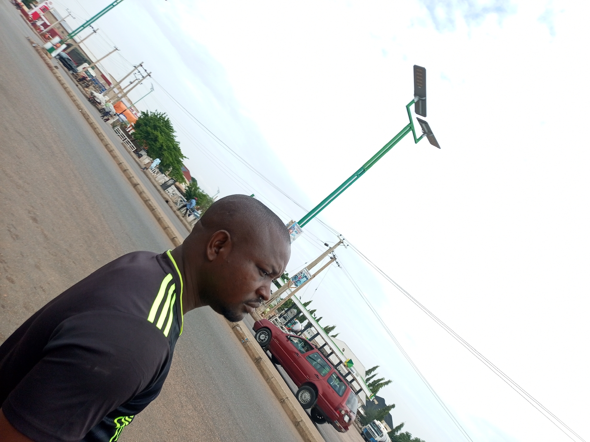 An uncaptioned, waist-up, medium shot shows a dark-skinned man with a shaved head, beard, and a black t-shirt with neon green stripes on the sleeves looking down and to his right. He is standing on a paved road, with power lines and streetlights visible in the background. A red SUV and a white truck are parked on the side of the road. The scene appears to be in an urban area, possibly Gombe, Nigeria, based on the style of the streetlights and the general environment.