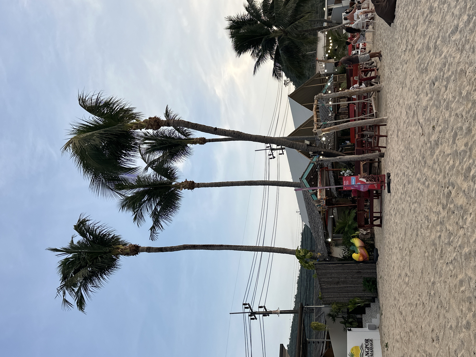 The image shows a beachfront scene in the Koh Samui District of Thailand. In the foreground, there is a sandy beach with a few chairs and tables set up, suggesting an outdoor dining or seating area. Several palm trees are prominent, with lush green fronds reaching towards the sky. A small structure, likely a hut or cafe, with a thatched roof and open sides, is visible behind the tables and chairs, decorated with string lights. Further back, there are more buildings and electrical poles with power lines. A sign on the left reads "ANGKOR MASSAGE". Several people are scattered around the area, some seated at tables and others standing. The sky is partly cloudy with a soft, diffused light, indicating it might be late afternoon or early evening. The overall atmosphere appears relaxed and tropical.