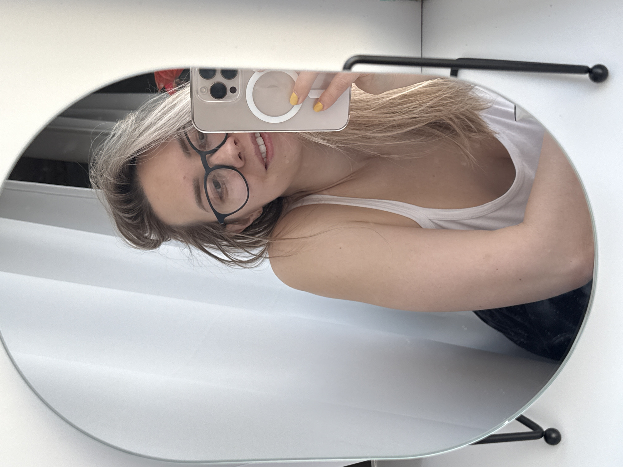 A woman is taking a selfie in a round mirror with a black frame and stand. She is holding a smartphone with a circular magnetic ring on the back in front of her face. The woman is wearing glasses and a white tank top. The mirror is placed against a white wall with a black towel rack. The location cannot be identified as Chaikovsky, Russia, or any specific city from the image.