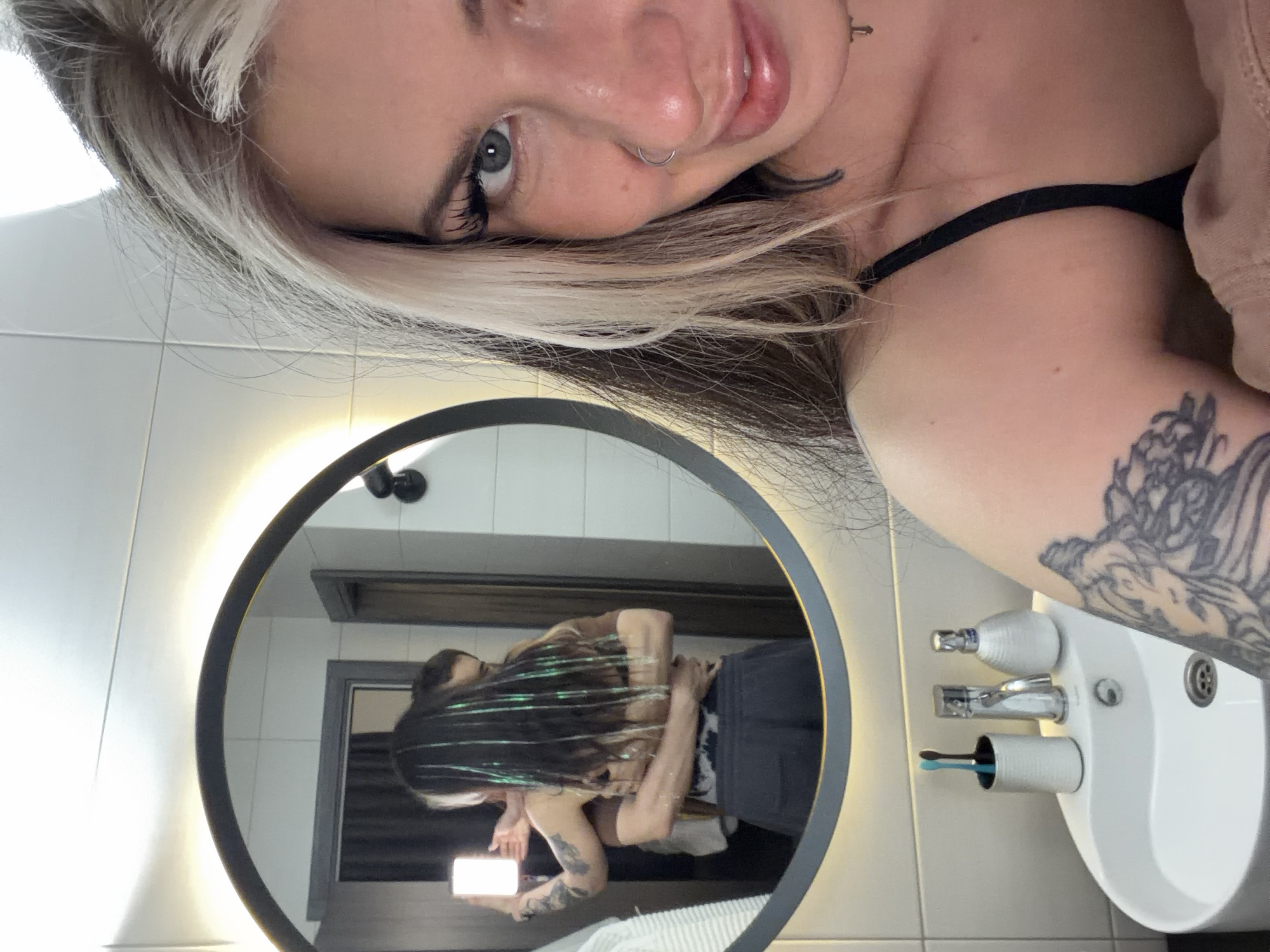 This selfie captures a young woman taking a photo in a modern bathroom. The image is oriented 90 degrees clockwise, showing the woman's face and upper body in the foreground, with her reflection visible in a large, round, backlit mirror.
The woman in the foreground has light blonde hair on top, layered over darker hair, and looks directly at the camera with clear blue-grey eyes. She has long eyelashes, a silver nose ring on her left nostril, and a relaxed, subtle smile with glossy lips. She is wearing a black strap top, possibly a bralette, with a light brown hooded garment partially visible. A detailed tattoo with an intricate design covers part of her right upper arm.
Her reflection in the mirror shows her holding a smartphone in her left hand, which is aimed at the mirror, confirming she is taking the photo. Her long, dark hair is adorned with distinctive, shiny green or blue strands, possibly tinsel or hair extensions. She appears to be wearing a light-colored top and dark pants or shorts. A tattoo is visible on her left arm in the reflection.
The bathroom setting includes a white ceramic sink with modern chrome fixtures, a soap dispenser, and a holder containing two toothbrushes (one blue, one black). The walls are light-colored, possibly tiled, and a dark-framed doorway is visible in the mirror's reflection, suggesting another room. The mirror itself has a dark frame and features a warm, circular backlighting effect. The scene conveys a casual, well-lit indoor environment, consistent with a private bathroom. Based on the provided context, the location is the Palilula Urban Municipality, Serbia. No visible text is present in the image.