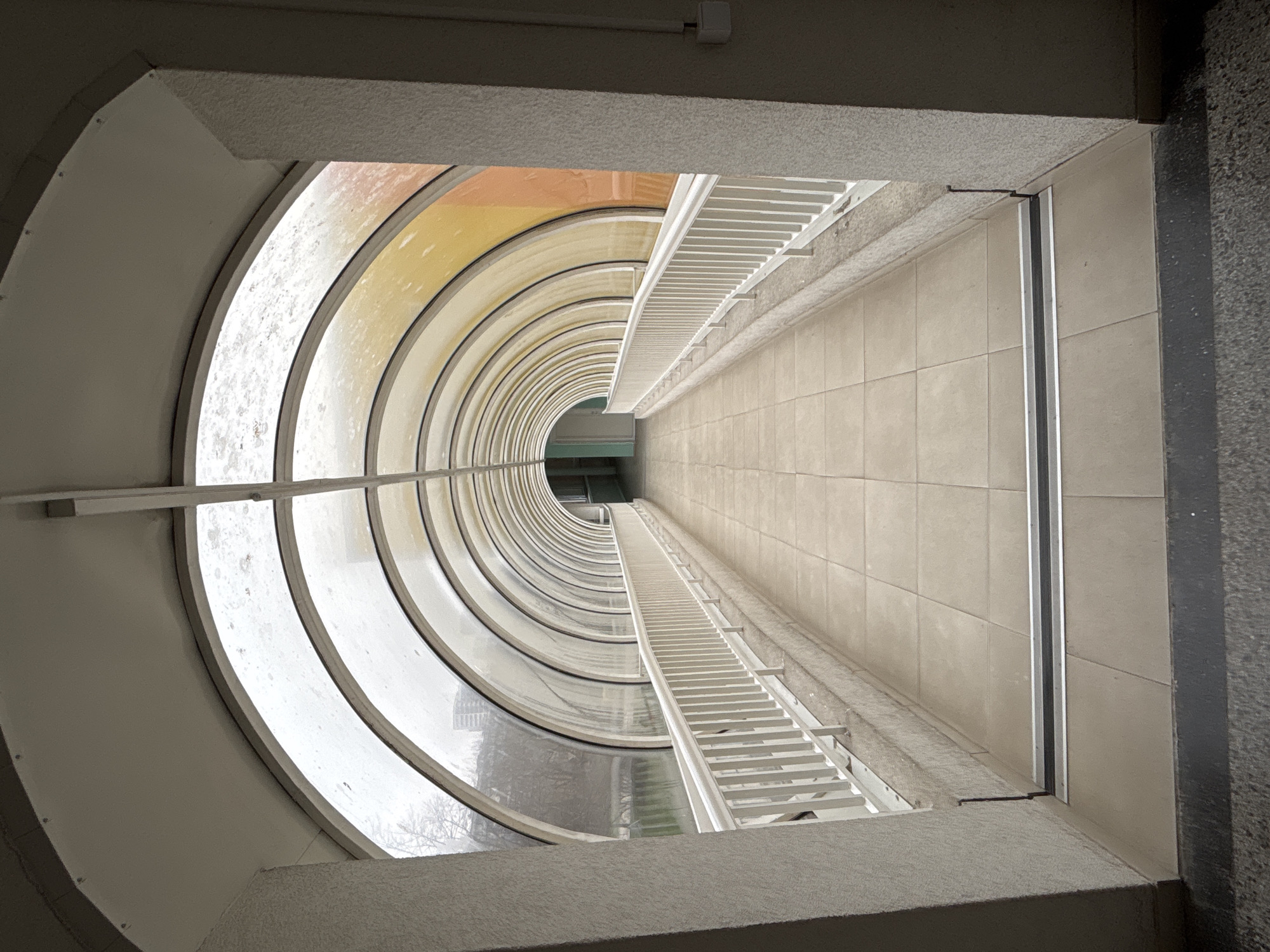 The image depicts an upward-looking perspective through a rectangular opening, revealing a cylindrical architectural feature. The cylindrical structure appears to be a stairwell or internal courtyard with curved windows or segments forming the exterior. The windows show variations in color, ranging from clear to yellow and orange hues. A white railing runs along one side of the rectangular opening and extends into the cylindrical structure. The walls inside the cylinder are tiled with a light-colored material. The rectangular opening is framed by concrete or plaster. At the top of the frame is a white electrical outlet box.
There are no people, subjects, objects, actions, or interactions visible. The environment is an indoor architectural space with natural light coming through the curved windows.
The location is not indicated in the image and cannot be determined.