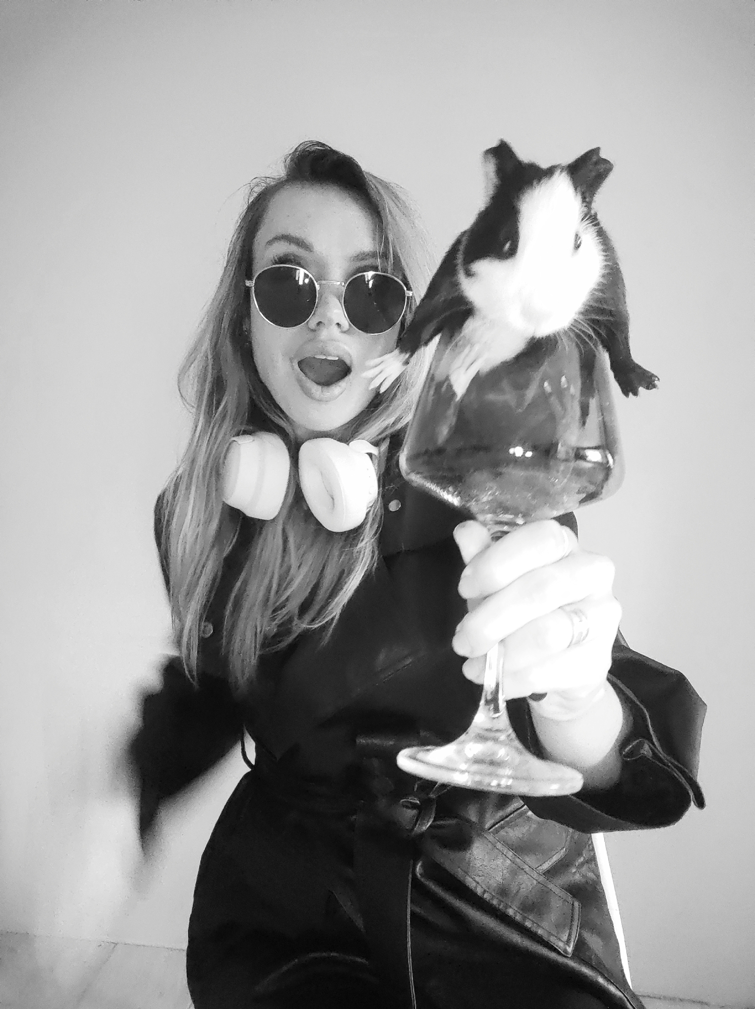 A black and white medium shot depicts a woman holding a large wine glass with a guinea pig perched on top. The woman wears round sunglasses, a dark belted jacket, and headphones around her neck. Her mouth is open in a surprised expression. The guinea pig is black and white. The background is a plain wall. It is not possible to determine the location in Votkinsk, Russia from the image.