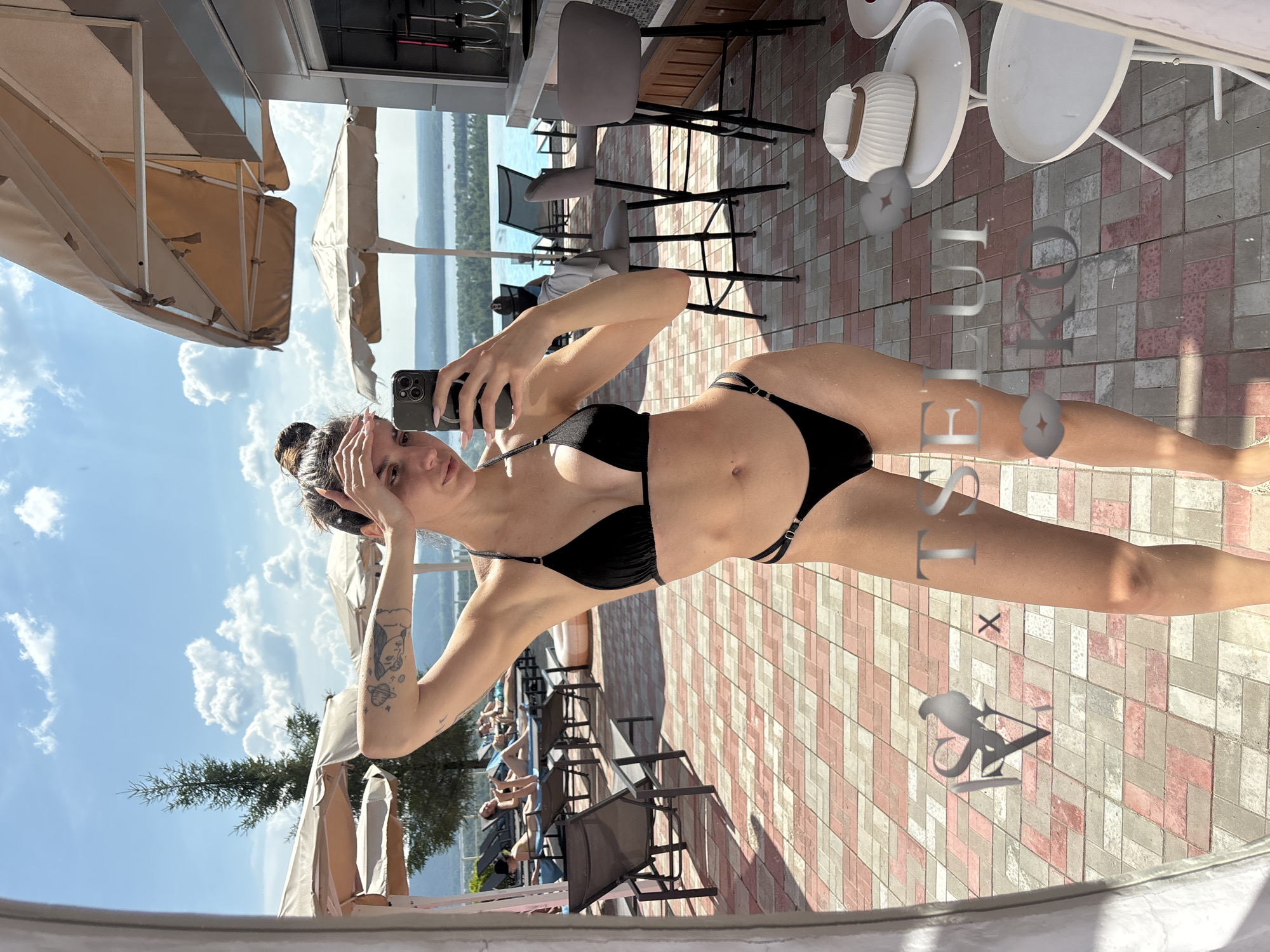 The image shows a young woman in a black bikini taking a selfie in a mirror. She is standing on a tiled patio with lounge chairs and umbrellas in the background. There is a body of water visible in the distance, possibly a river or lake, with a city skyline on the opposite shore. The overall scene suggests a resort or hotel setting. The woman has a tattoo on her left arm and her hair is pulled up in a bun. She is holding a smartphone in her right hand and has her left hand on her forehead. The lighting indicates it is a sunny day. Based on the provided information, the location is the city of Novokuybyshevsk, Russia.