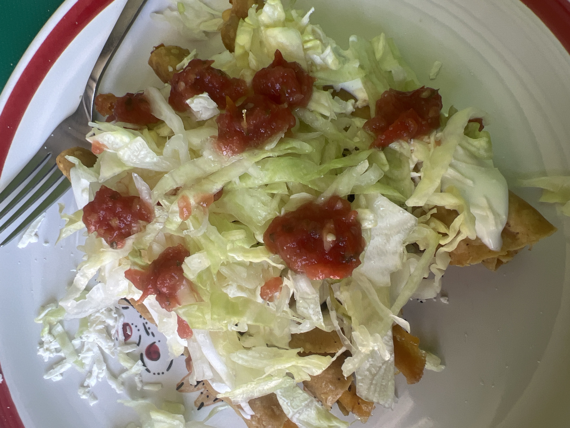 The image shows a plate of food, likely tostadas, with shredded lettuce and salsa on top. A fork rests on the plate next to the food. The plate is white with a red rim. Part of a green surface is visible in the background. The location is unknown.