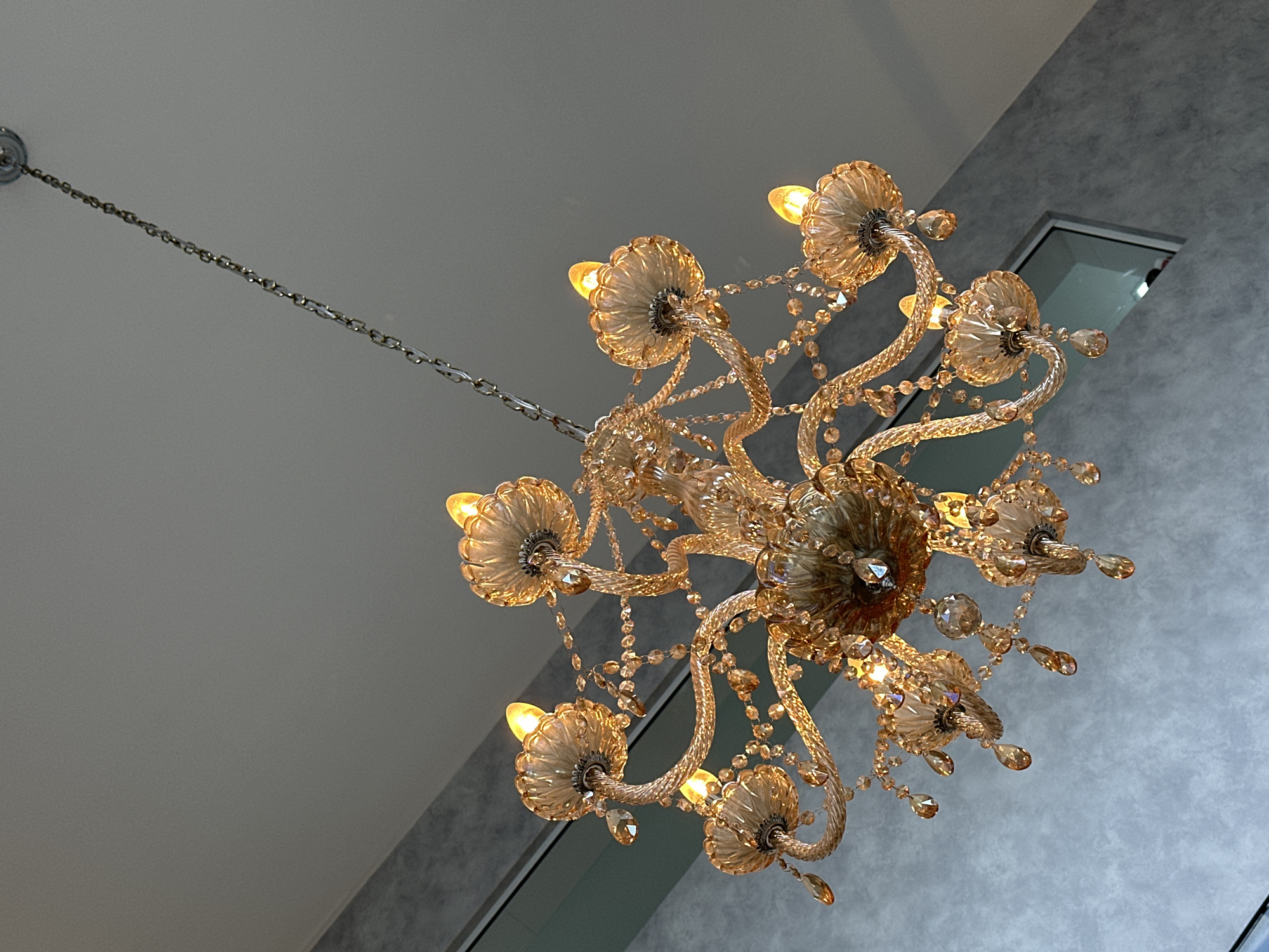 A chandelier hangs from a ceiling in what appears to be an indoor space with textured grey walls. The chandelier is ornate, with multiple arms made of amber-colored glass, adorned with crystal droplets and flower-shaped lamp holders. Each lamp holder contains a lit, amber-colored bulb, casting a warm glow. The chandelier is suspended by a silver chain. The background consists of a grey ceiling and a section of a grey wall with a subtle textured pattern, along with a window with a muted reflection. There are no people visible in the image. The setting does not provide any specific visual cues to identify it as being located in Florianópolis, Brazil.