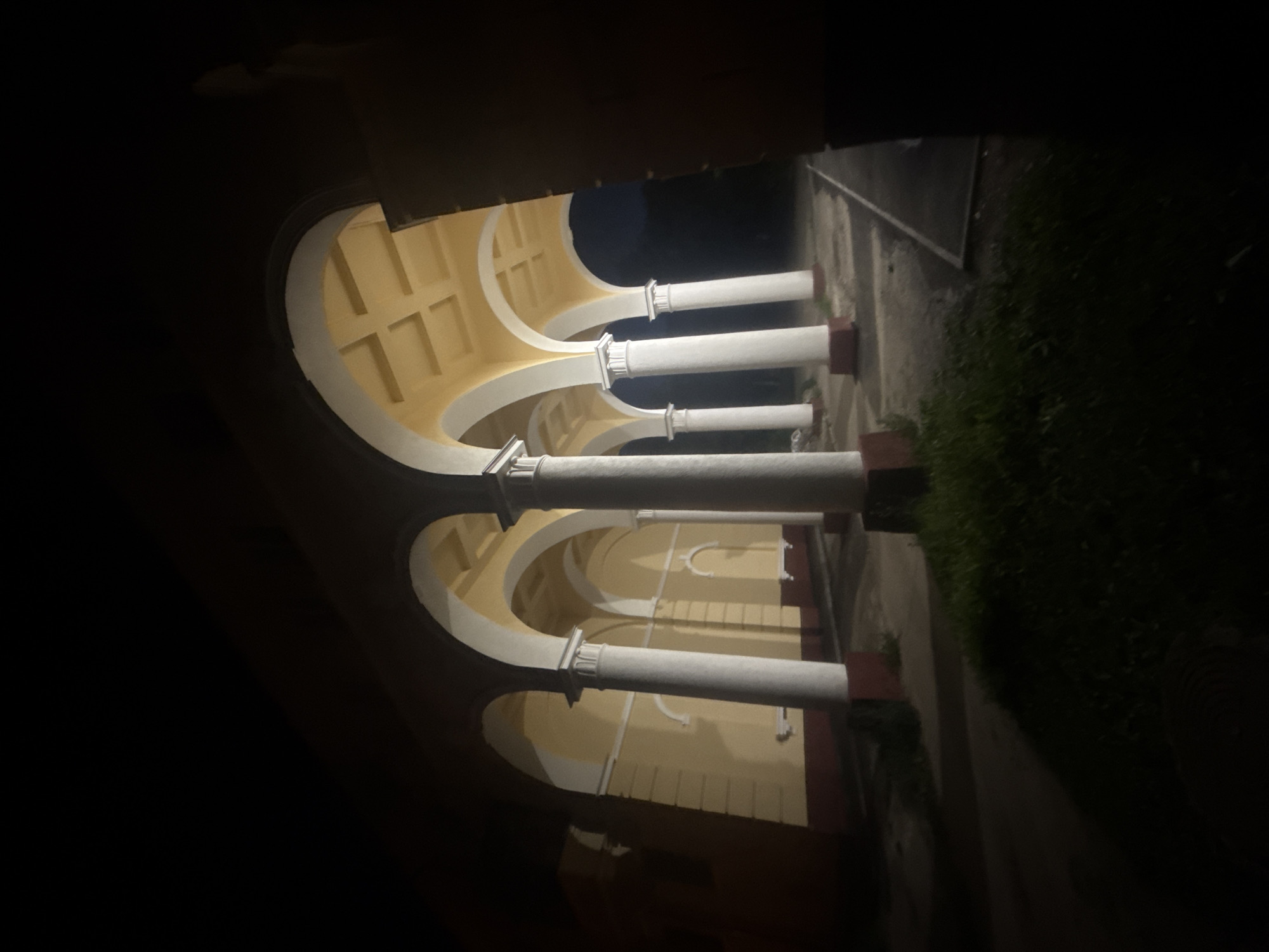 The image depicts a dimly lit architectural structure with a series of arches supported by white columns. The building appears to be yellow with a red base. The ceiling of the arched walkway has a coffered design. The ground in front of the structure is paved, and there is some vegetation visible. The scene is set at night, with the architecture illuminated by an unseen light source that casts shadows. There are no people or animals present in the image. The scene is located in Magnitogorsk, Russia.