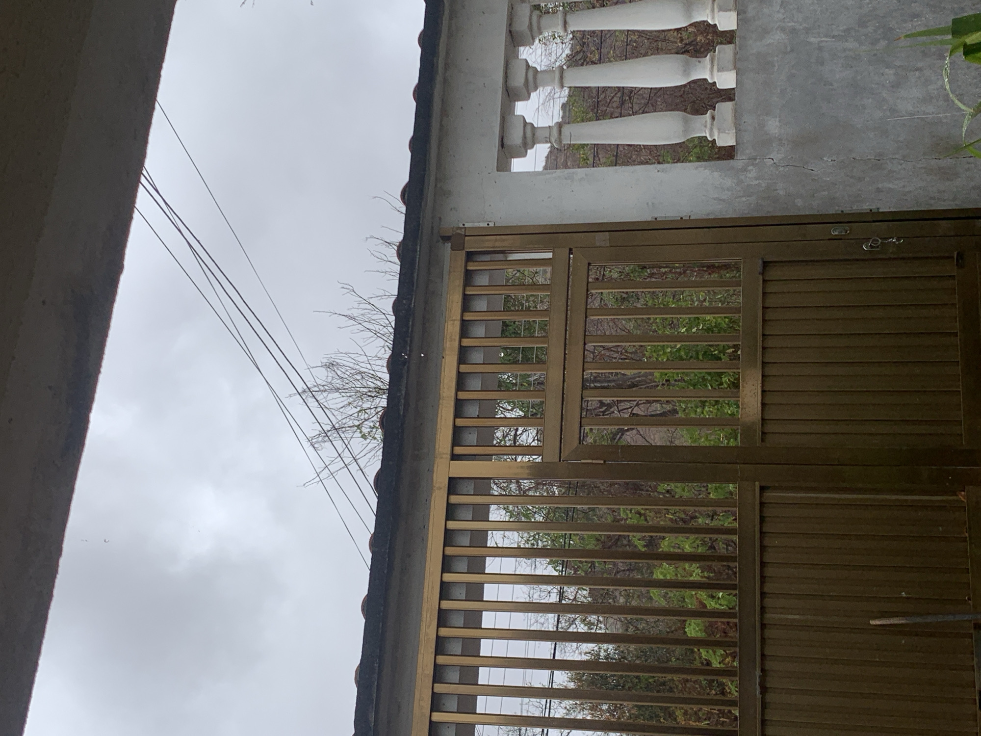 The image presents an upward-angled view of a building exterior, with a cloudy sky visible in the background. The structure includes a concrete balcony with white balusters, a gate constructed of gold metal bars, and a tiled roof edge. Wires are running in the foreground. The scene appears to be in Santa Ana, Venezuela.