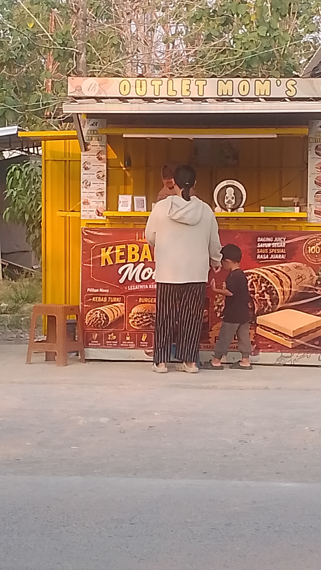 The image depicts a street-level view of an "OUTLET MOM'S" food stand located in Karanglangu, Indonesia. The stand is painted bright yellow and features a large red banner advertising "KEB...Mo" which appears to be a food item. The banner displays images of a kebab and a burger. Behind the counter, at least one person is visible, likely an employee. In the foreground, two people are interacting with the stand: an adult woman wearing a light-colored hooded sweatshirt and striped pants, and a young boy in a dark t-shirt and grey pants. The boy is gesturing towards the stand. To the left of the stand, a small brown plastic stool is placed on the ground. The environment appears to be outdoors, with trees and vegetation visible in the background. The ground is paved with asphalt.