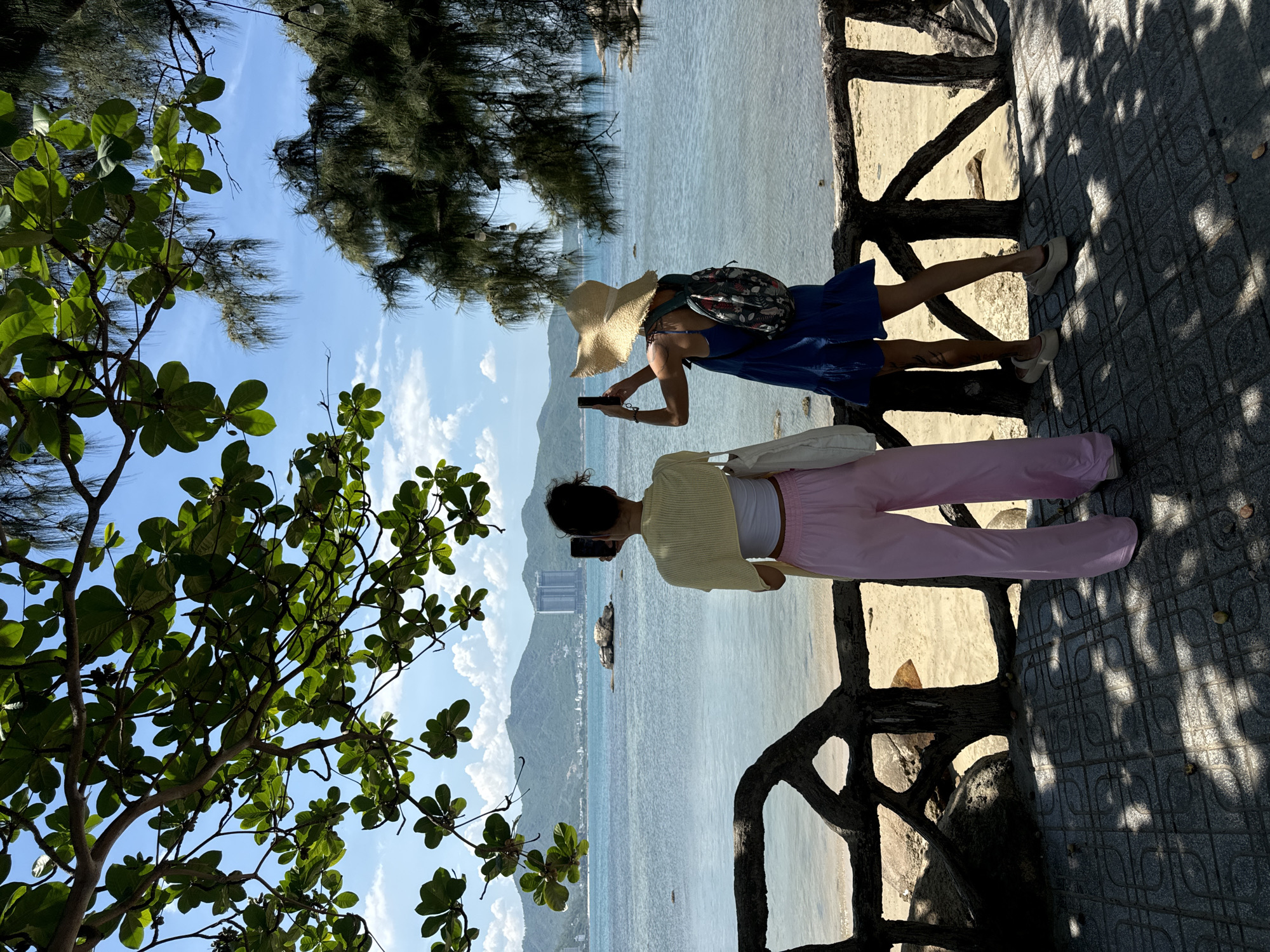 Two women are standing on a tiled pathway overlooking a beach and the sea in Nha Trang, Vietnam. The woman on the left, facing away from the camera, is wearing a yellow cardigan over a white top and pink wide-leg pants. She holds a black smartphone in her right hand, taking a photo. She carries a white tote bag over her left shoulder. The woman on the right, also facing away, wears a blue dress and a straw hat. She has a backpack on and is holding a black smartphone in her right hand, taking a photo. The pathway is bordered by a rustic wooden fence with abstract cutouts. Behind the fence, a sandy beach leads to the calm, blue sea. In the distance, a coastline with green mountains and buildings is visible under a partly cloudy sky. Lush green foliage from a tree hangs overhead, casting dappled shadows on the pathway.