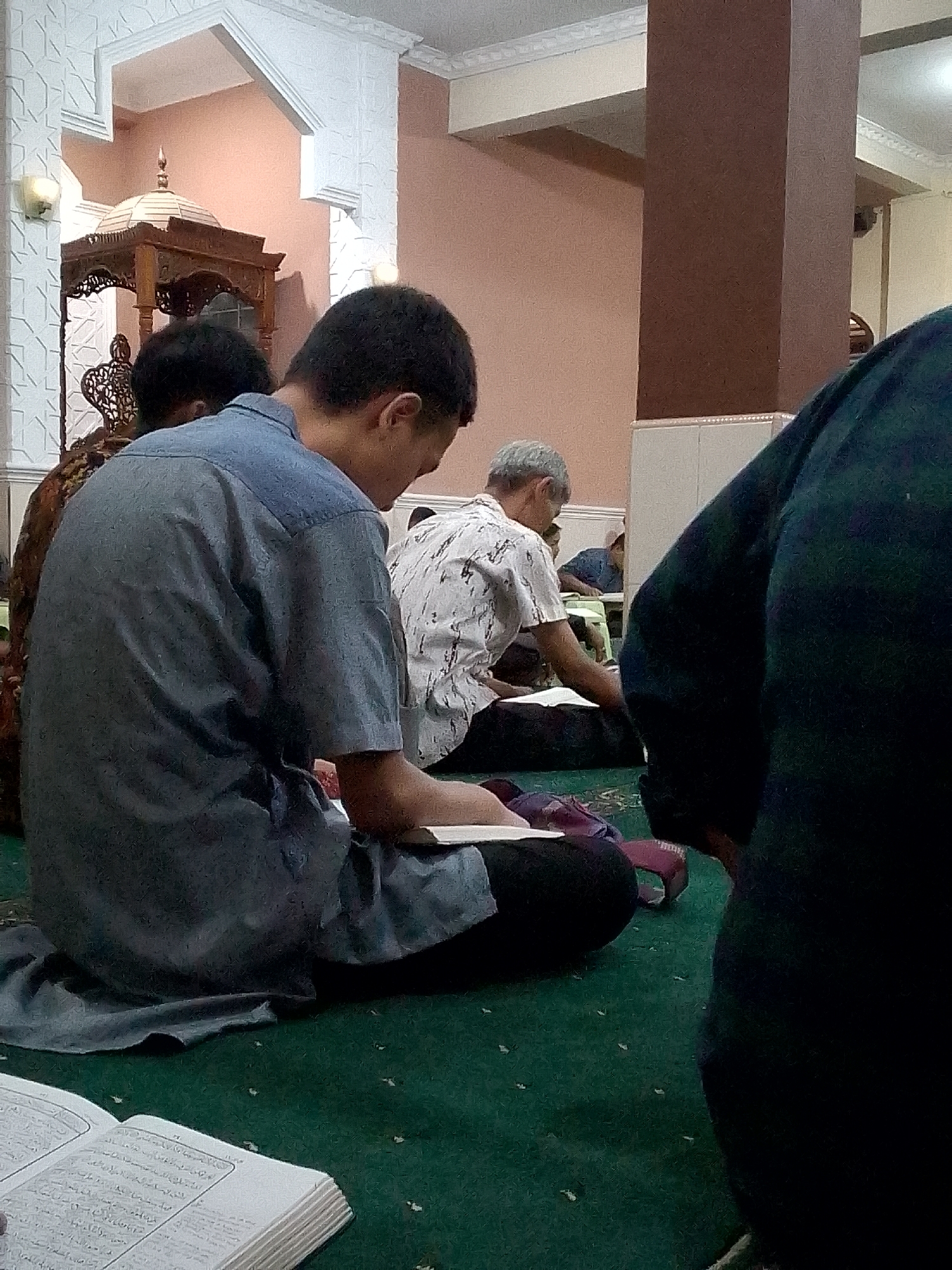In Kertajaya, Indonesia, a group of men are seen engaged in religious activities within a mosque. The foreground is dominated by a young man wearing a blue shirt, seated on a green carpet, engrossed in reading a book. To his left, a person in a patterned garment is also reading. Further back, an older man with gray hair, wearing a white shirt, is looking down at a book. Another individual, partially visible, is seated behind him. The environment is characterized by ornate architectural details, including a white arched structure and a wooden altar-like structure with a dome. The lighting suggests an indoor setting, possibly during prayer time.