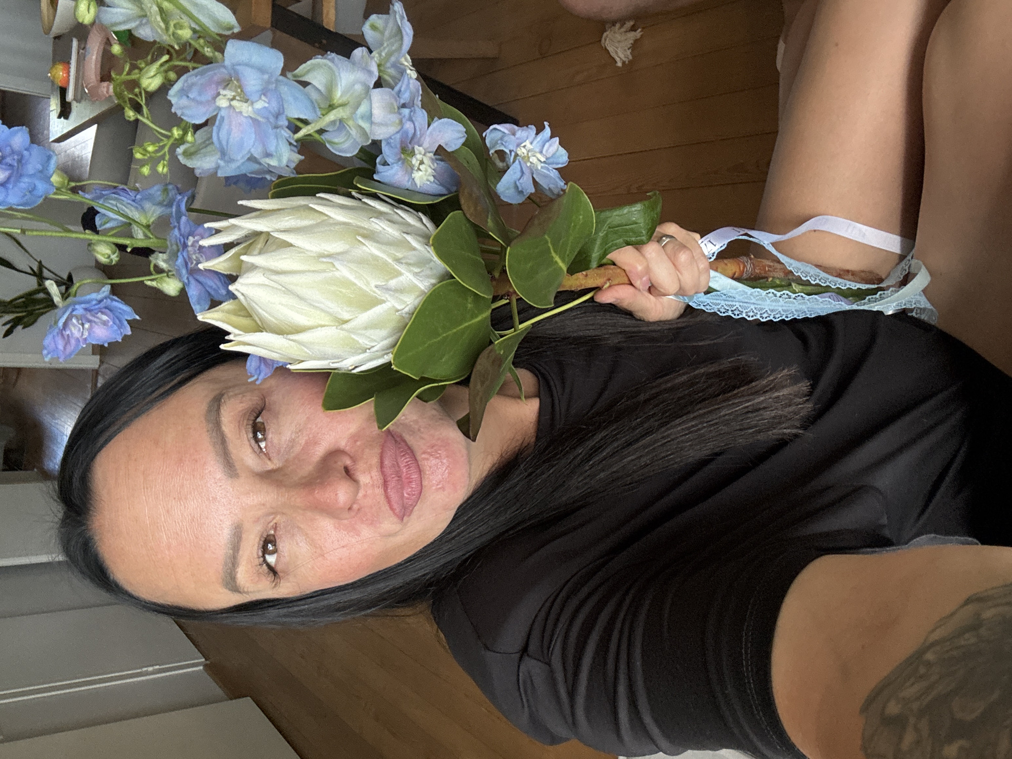 A selfie is taken by a woman with dark hair and brown eyes, who is looking directly at the camera with her lips pursed. She is holding a bouquet of flowers, which includes a large white protea and several blue delphiniums. She is wearing a black t-shirt. The background shows a wooden floor and a glimpse of a table with some items on it. The scene is indoors.