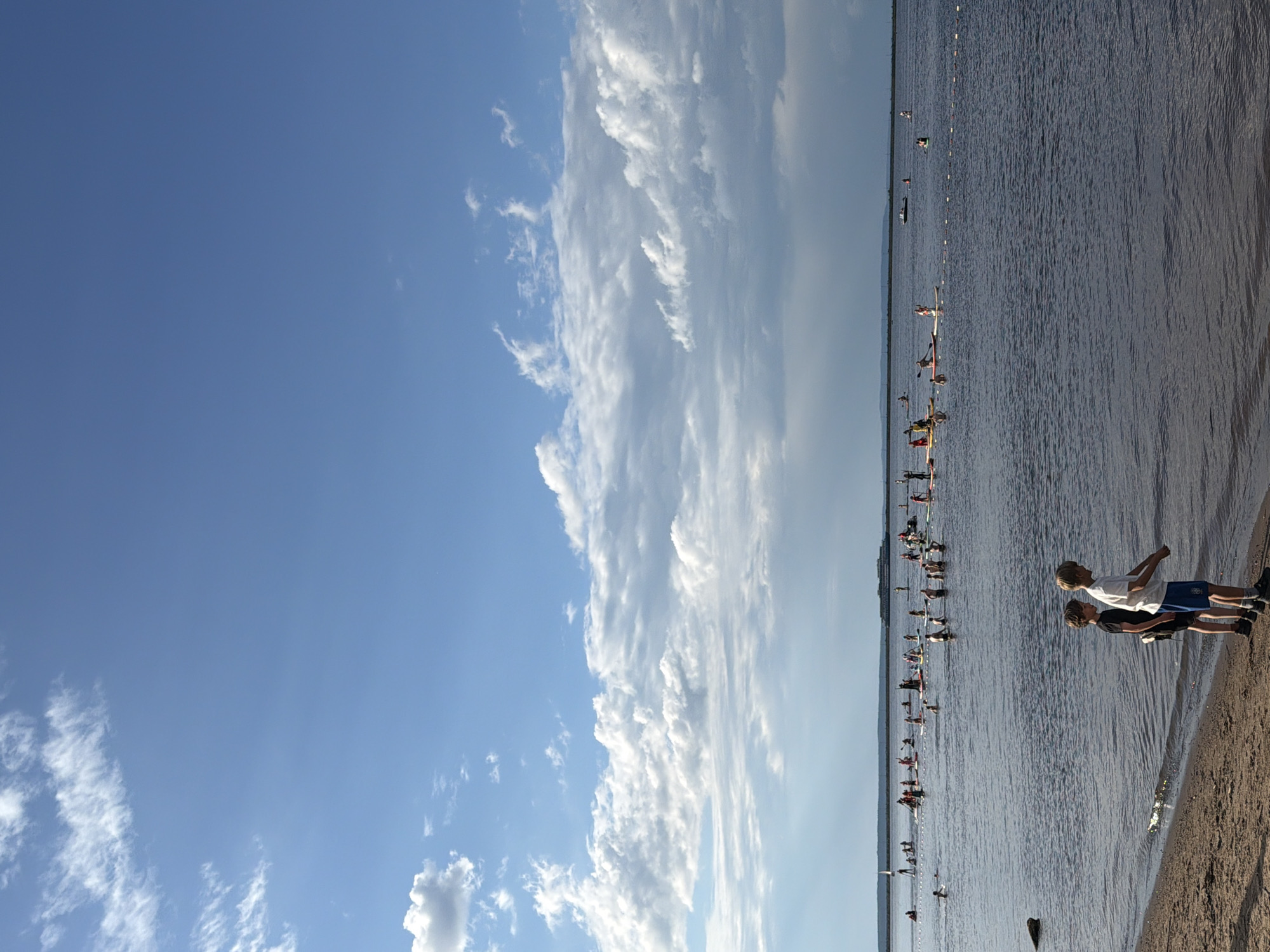 This media file depicts a sunny day at a body of water, likely a lake or river, with many people engaged in paddleboarding. The scene is viewed from the shore, looking out across the water towards a distant treeline or buildings. The sky is a clear blue with some scattered white clouds, suggesting fair weather and likely daytime.
The foreground features a sandy shore where two young boys are standing, looking out at the water. They appear to be observing the activity. The water is calm with gentle ripples, reflecting the sky. A multitude of paddleboarders are spread across the water, some closer to the shore and others further out. They are spread out in a long line, indicating a potential event or gathering.
The overall mood is one of leisure and recreation. The bright sunlight and clear sky contribute to a positive and pleasant atmosphere. The distant landscape is hazy, possibly due to the distance or atmospheric conditions. There is no visible text in the image. The orientation of the image suggests it was taken in a vertical format, and has been rotated. The context provided places this scene in Chaikovsky, Russia.