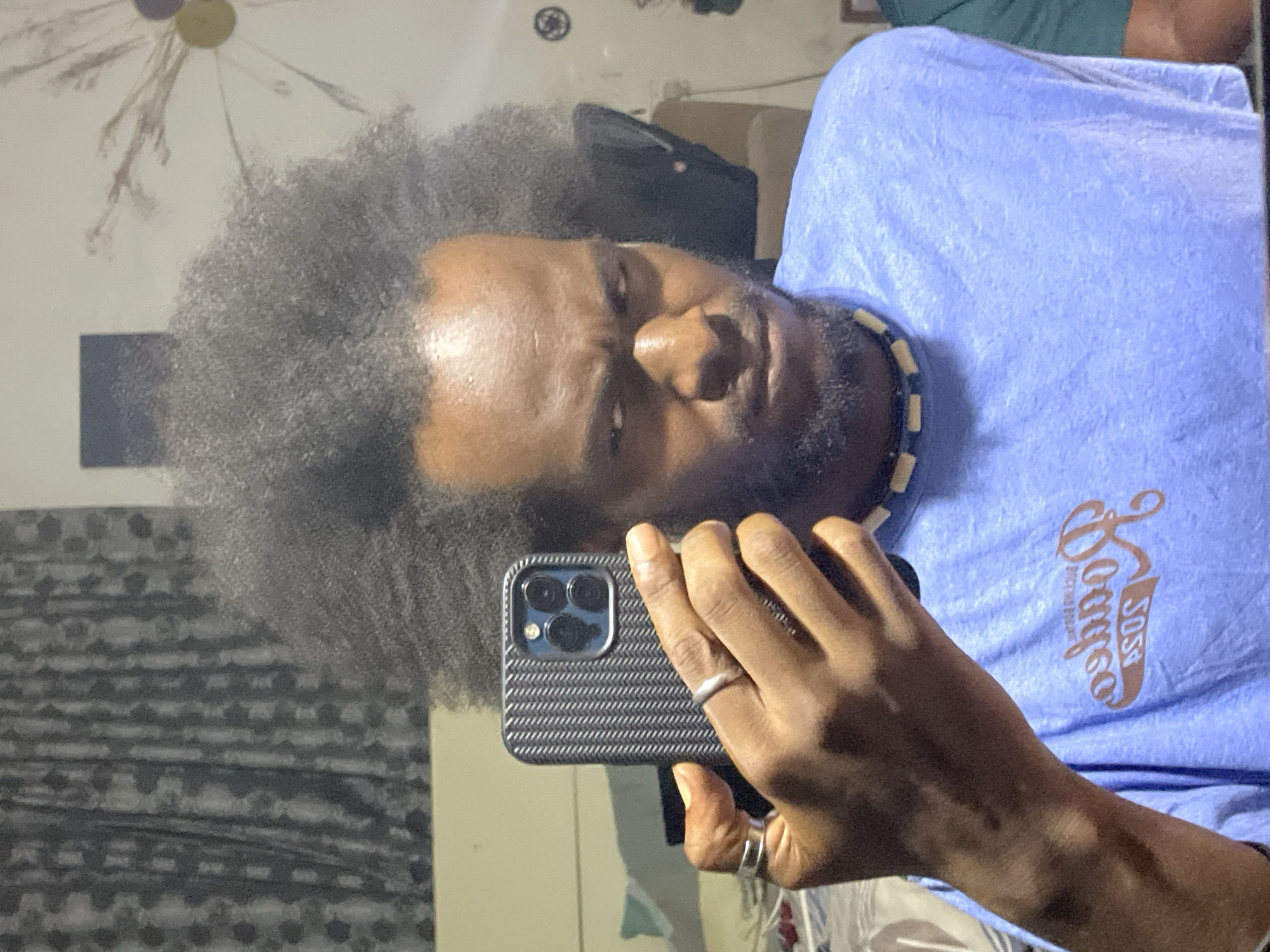 A medium shot shows a Black man taking a selfie with a phone. He has an afro hairstyle and is wearing a light blue t-shirt with "SOSA Lounge" printed in an elegant brown font. A necklace of light-colored cylindrical beads and dark flat circular beads is visible around his neck. The phone has a textured, dark-colored case, and he is wearing two rings, one on his ring finger and one on his pinky finger. The background includes a patterned curtain, a light-colored wall, and various decorative elements. The location is not identifiable as Kuchingoro, Nigeria, based on the visual details in the image.