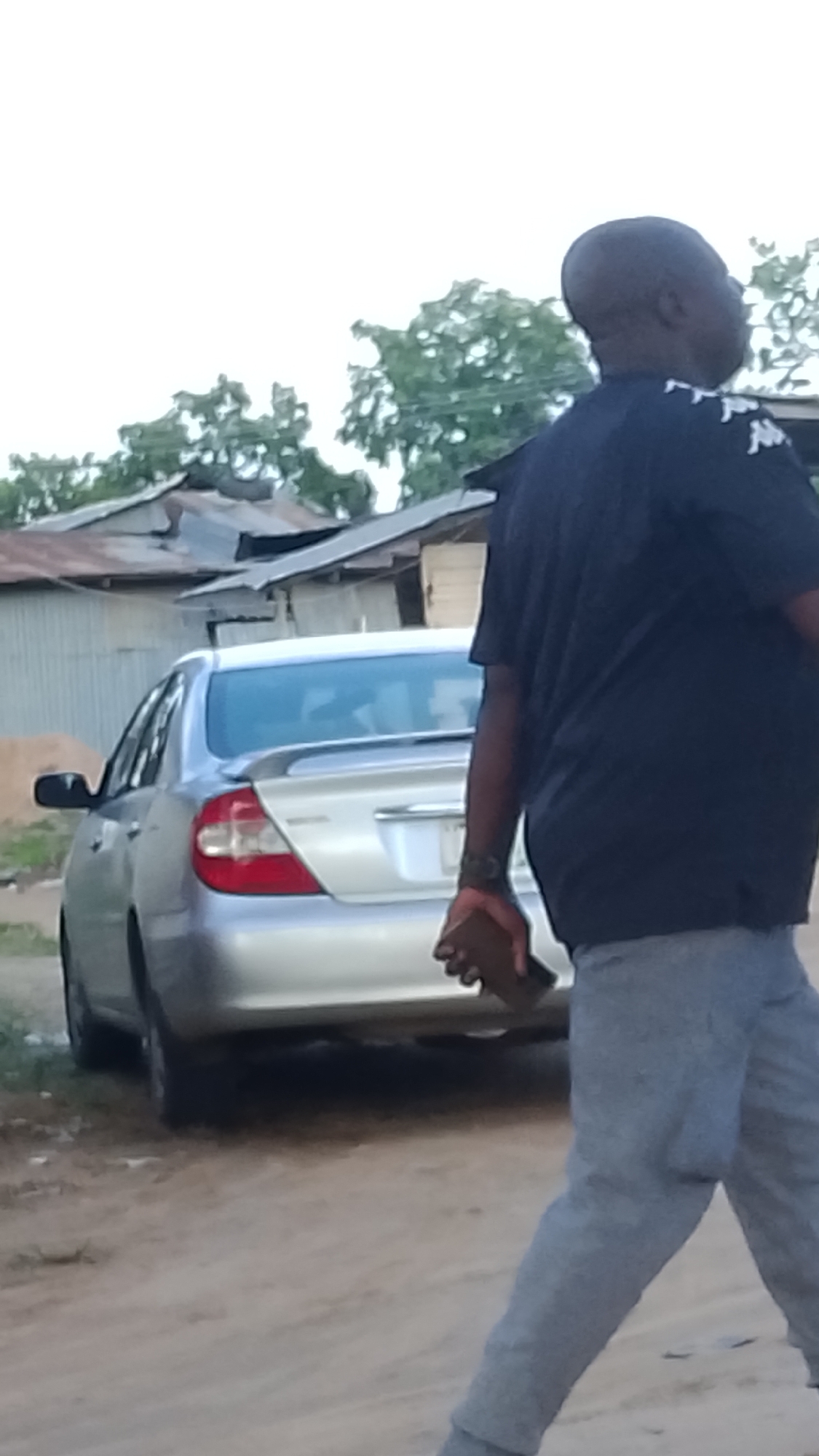 A man, viewed from behind, walks on a dirt road in Adankolo, Nigeria. He is wearing a dark blue t-shirt with white emblems on the shoulders and light gray sweatpants. He is holding a dark brown object in his right hand. Behind him, a silver sedan is parked on the side of the road, facing away from the viewer. In the background, there are several buildings with corrugated metal roofs, and trees. The sky is overcast.