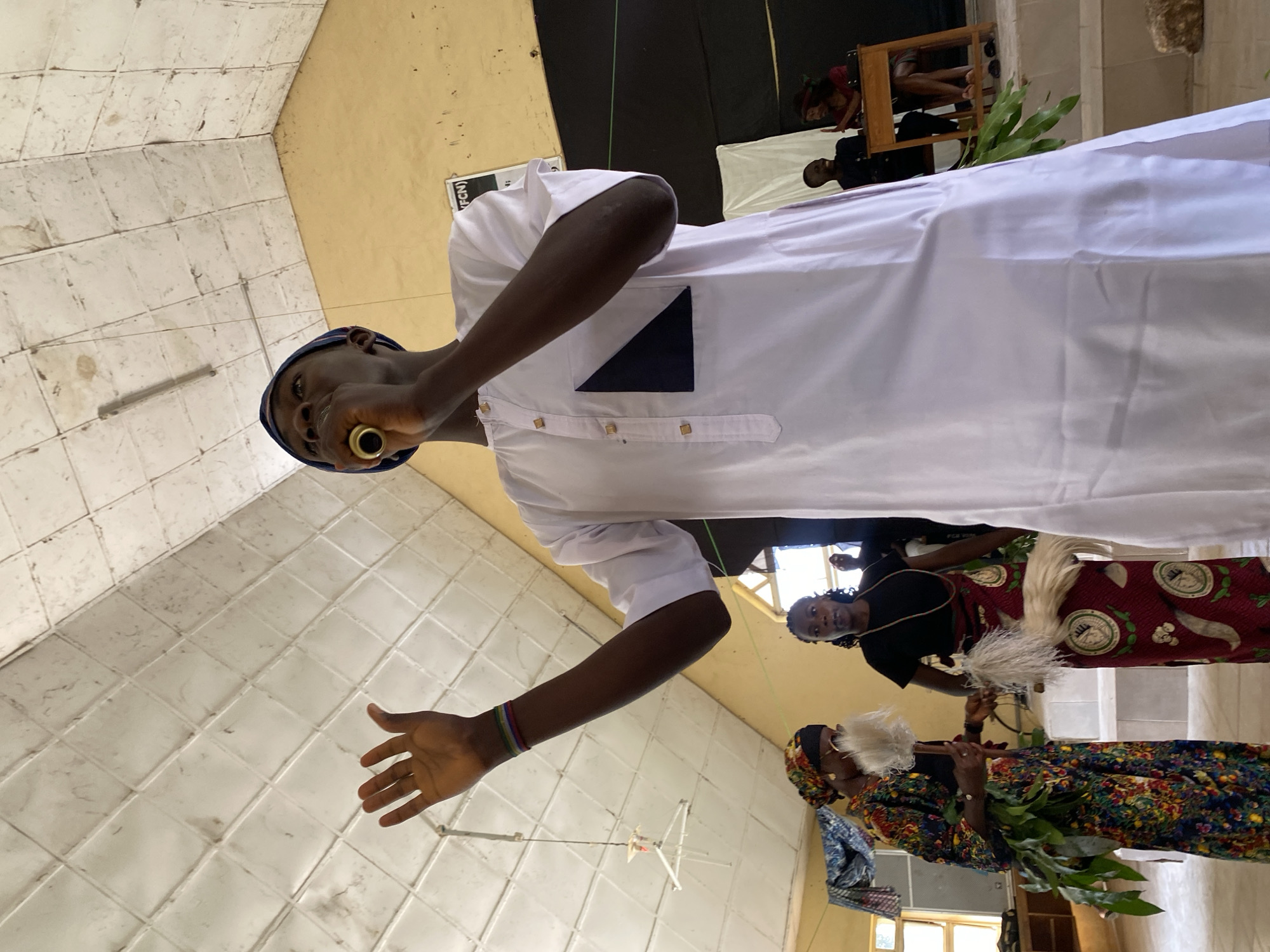 In Dutsen Kato, Nigeria, three individuals are captured in a photograph, with the perspective rotated by 90 degrees clockwise. The primary subject is a person in the foreground, dressed in a white, long-sleeved garment with gold buttons and a dark blue geometric accent on the left chest. They wear a dark blue cap and a gold ring in their lower lip. Their left arm is extended, palm open, and they appear to be speaking or singing into a golden object held near their mouth.
Behind this central figure, two women are visible. The woman on the left is wearing a black top and a patterned skirt featuring red and white motifs. She is looking towards the viewer. The woman on the right is dressed in a colorful, patterned attire, including a headscarf, and holds a whisk-like object in her right hand. She is also holding what appears to be greenery or branches in her left hand.
The setting seems to be indoors, with tiled walls and a yellow ceiling. In the background, a white cloth is draped, and a wooden chair with a person seated on it is partially visible. There is also a sign with some text, but it is mostly obscured. The overall lighting suggests daytime. The expressions of the people are difficult to ascertain with certainty due to the image rotation and resolution, but the scene appears to be part of a cultural gathering or ceremony.