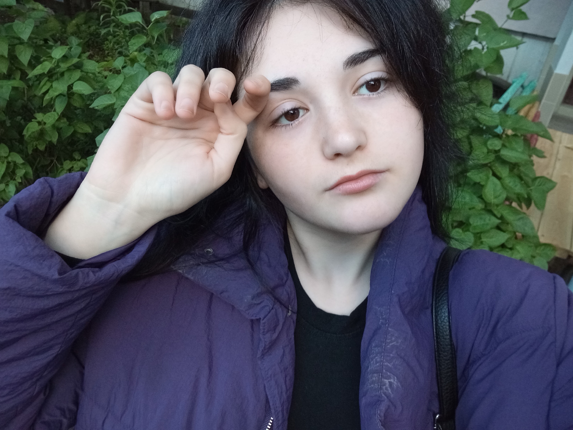A close-up, head-and-shoulders portrait features a young woman outdoors in Poltava, Ukraine. She has long, dark hair, dark brown eyes, and prominent, thick eyebrows, with a fair complexion. She looks directly at the camera with a neutral and composed expression. Her left hand is raised, with her fingers gently touching her left eyebrow, creating a subtle gesture or appearing to shield her eyes.
She is wearing a dark purple or indigo jacket, possibly a windbreaker or lightweight puffer style, which exhibits a slightly wrinkled texture. Beneath it, a black t-shirt is visible around her neckline. A black strap, likely from a shoulder bag, crosses over her right shoulder.
The background consists of dense, lush green foliage, indicating an outdoor setting such as a garden, park, or a green area within a residential space. On the far right, behind the greenery, there are faint hints of a light-colored building or structure. The natural, soft lighting suggests the photo was taken during the daytime. There is no visible text in the image.