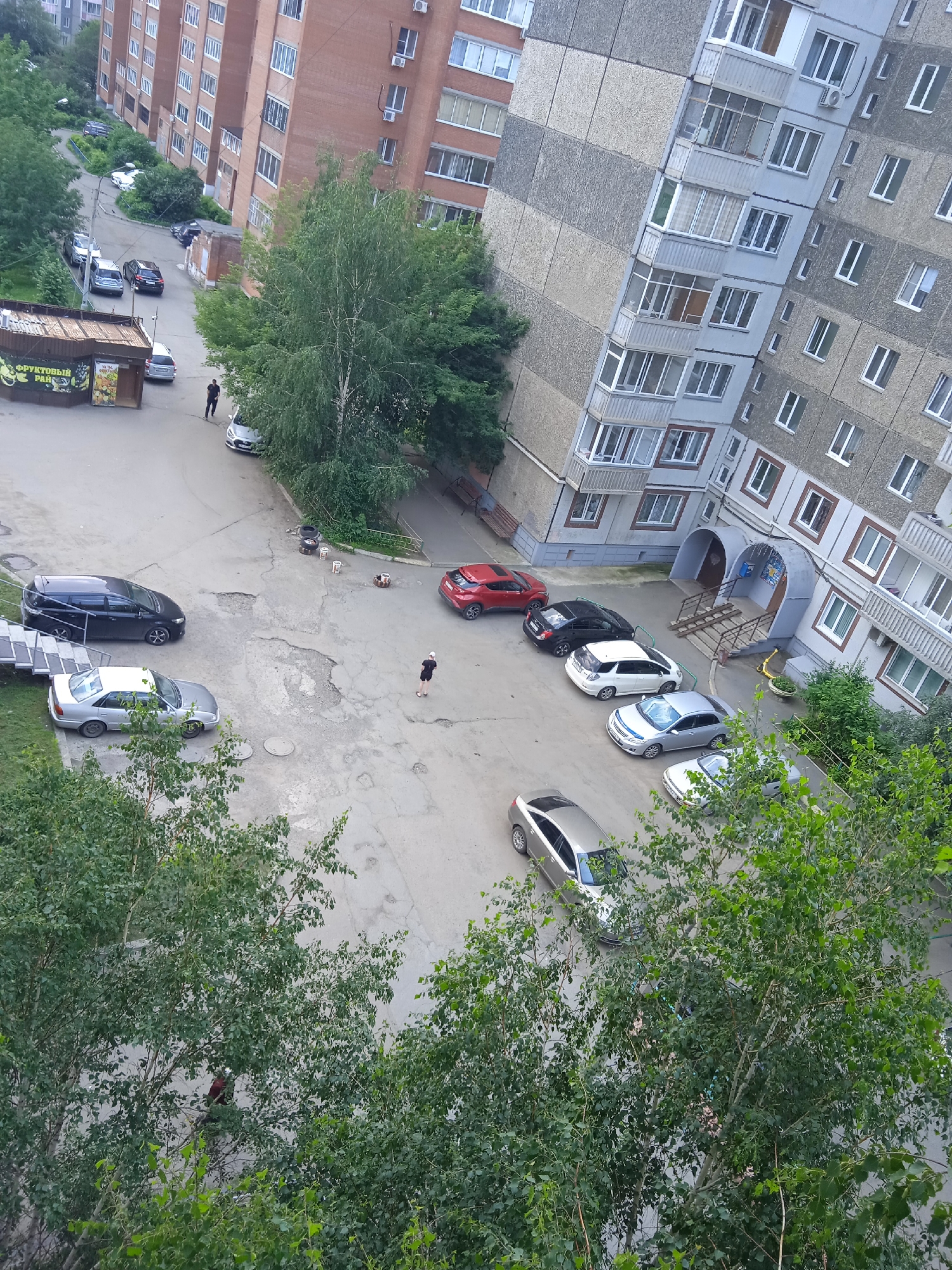This is a high-angle, medium shot of a residential courtyard in Krasnoyarsk, Russia. The scene is dominated by a large, multi-story, grey and beige apartment building with numerous windows and balconies. Adjacent to it is a reddish-brown brick apartment building. The courtyard is paved with asphalt and shows signs of wear and tear with several large potholes.
Several cars are parked in the courtyard: a black minivan, a silver sedan, a white hatchback, a black sedan, a red hatchback, and another silver sedan further down the road. Two people are visible: one individual, dressed in dark clothing, is walking across the paved area, and another person, wearing a white cap and dark clothing, is standing near a red car. A small kiosk with signage is visible on the left side of the image, possibly a convenience store. Lush green trees with dense foliage surround the paved area and partially obscure the buildings. The sky is overcast, suggesting a cloudy day.