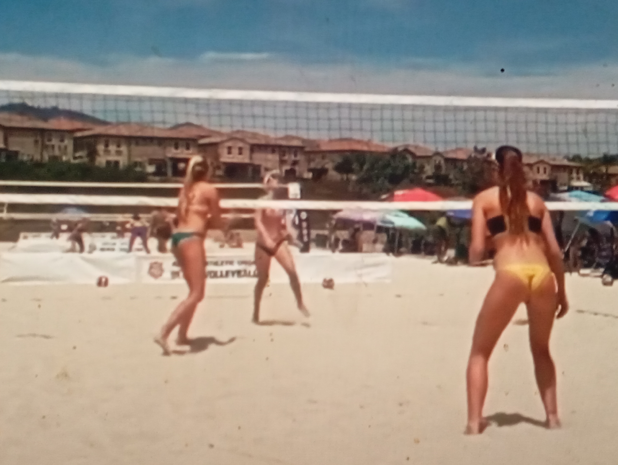 The image depicts a beach volleyball game. Three women are visible on a sandy court, with a volleyball net strung across the middle. One woman, in the foreground and facing away from the camera, wears a black bikini. To her left, another woman in a green bikini is in motion, suggesting she is playing. Further left, a third woman, also in a bikini, appears to be observing the game or waiting for action. In the background, rows of houses with tiled roofs are visible, along with some trees and umbrellas, indicating an outdoor recreational area. The sky is a clear blue with a few faint clouds. The scene appears to be a sunny day.