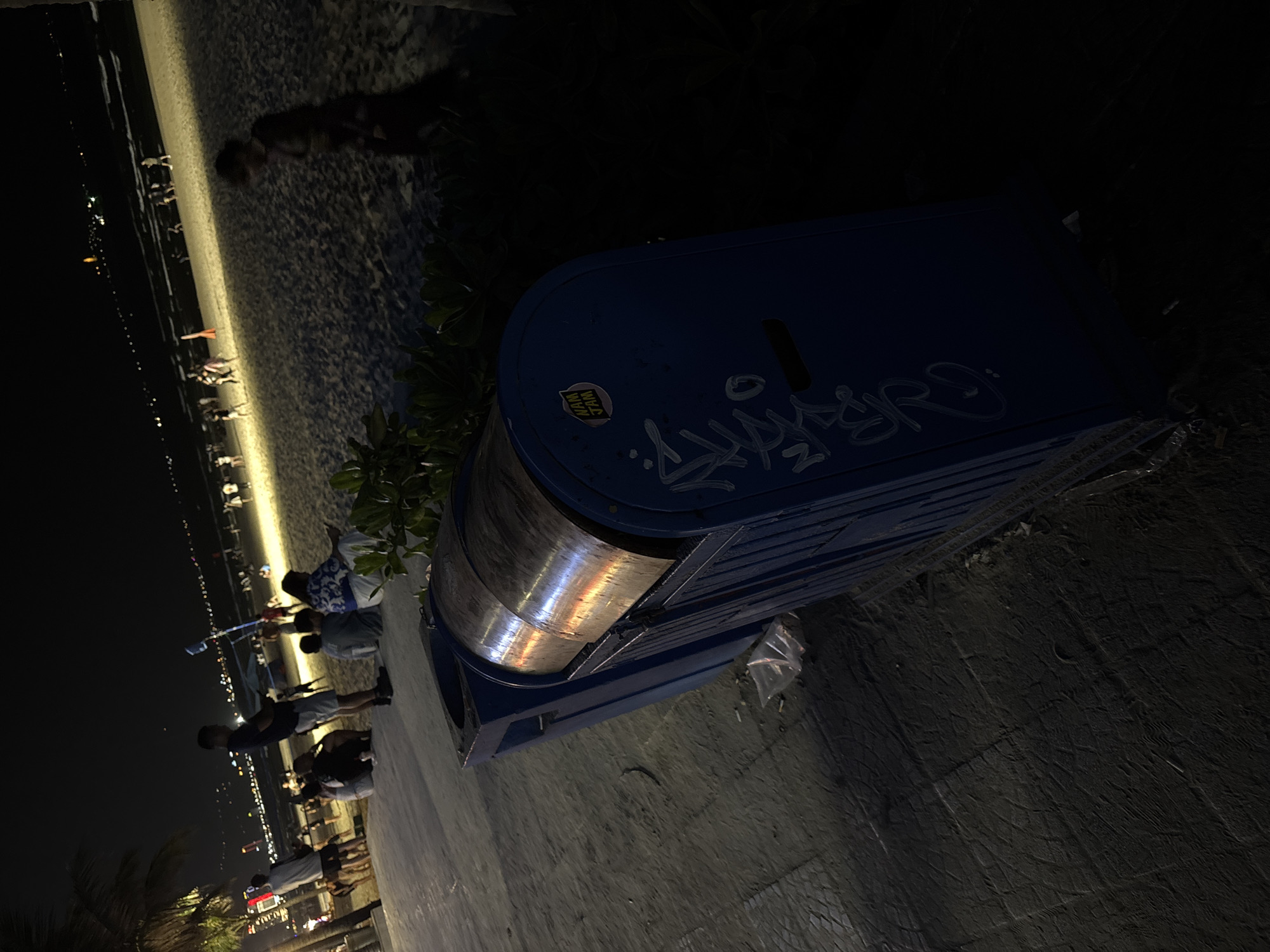 At night in Đà Nẵng, Vietnam, a blue trash can with graffiti and a sticker is placed on a paved area. The background shows a beach with people and city lights.