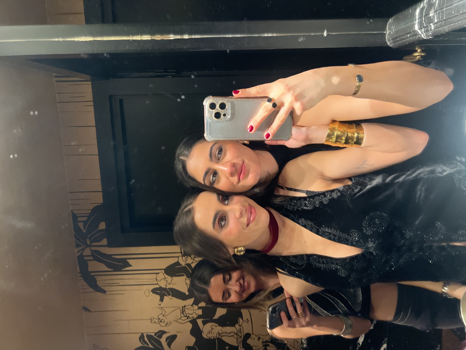 Three women are taking a mirror selfie. The top woman holds an iPhone, and wears a black top. The woman below wears a black dress with a red choker necklace and gold earrings. The woman at the bottom wears black with silver accents, and holds a phone. The photo shows the mirror's reflection of the three women. The wall has black and tan wallpaper.