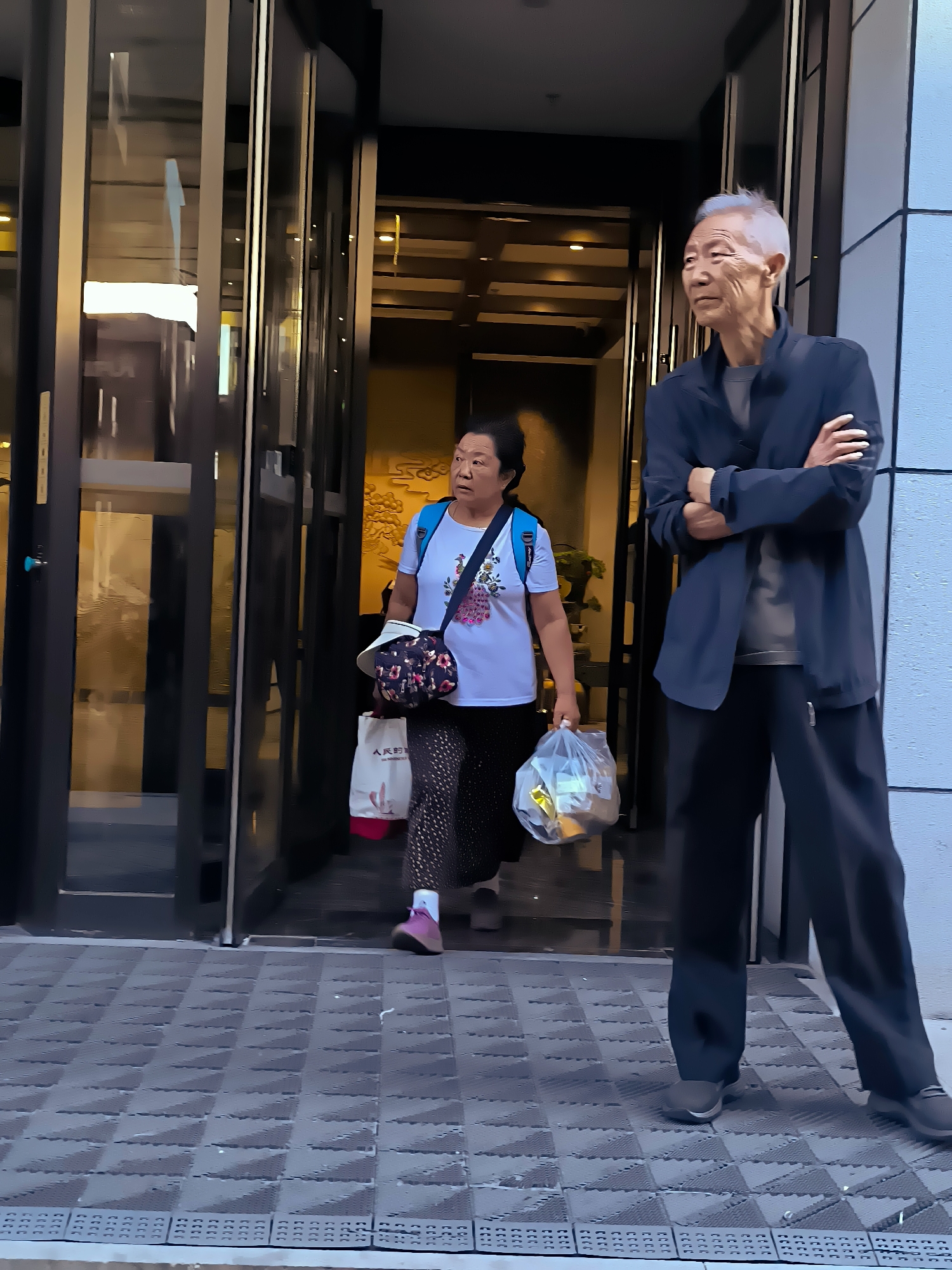 In Ürümqi, China, two individuals are positioned on a patterned outdoor tile walkway in front of a modern building with large glass doors. On the right, an elderly Asian man stands with his arms crossed, wearing a dark jacket over a grey t-shirt and dark pants. He has grey hair and is looking to his right. To his left, an elderly Asian woman walks out of the building. She is wearing a white t-shirt with a floral design, dark patterned pants, and pink shoes. She carries a white shopping bag with red accents in her left hand and a clear plastic bag filled with items in her right hand. A floral-patterned purse hangs from her shoulder. The building entrance features dark framing and glass panels reflecting the interior.