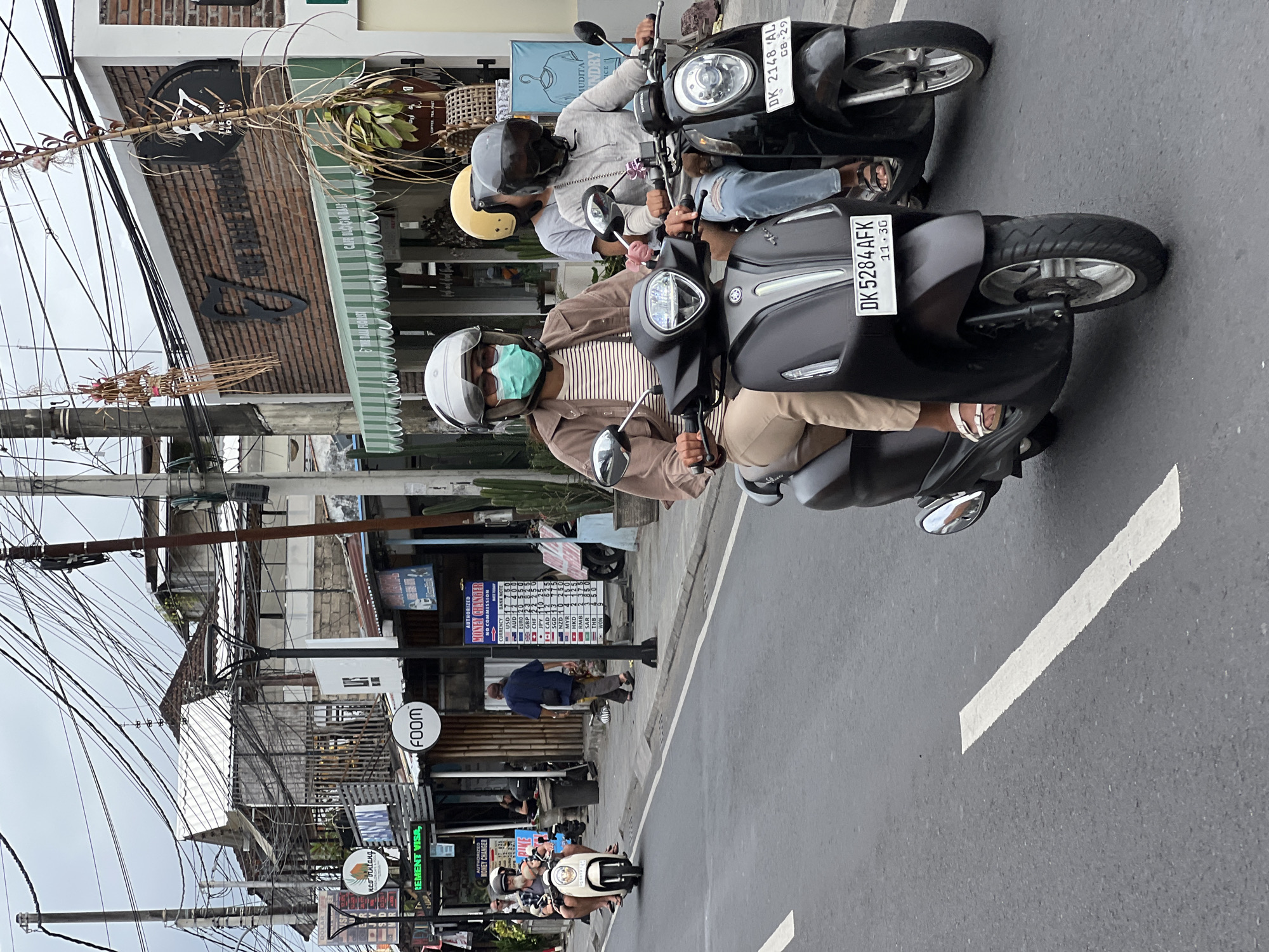 A medium shot captures a street scene in Denpasar, Indonesia, on a cloudy day. Three individuals on scooters are prominent in the foreground, navigating the asphalt road. The scooter closest to the camera is dark gray, ridden by a person wearing a light brown jacket over a striped shirt, beige pants, and white sandals. This person is also wearing a white helmet and a light blue face mask. Behind them, on a black scooter, are two individuals. The rider wears a light gray hoodie and blue jeans, with a black helmet. The person behind them, partially obscured, has a yellow helmet. Both scooters have visible license plates: the dark gray one reads "DK 5284 AFK 11.3G", and the black one reads "DK 2148 AL 08.29".
The background reveals a bustling street with various buildings, including shops and cafes. A prominent building on the right has a brick facade with the signage "THE BALI FLORIST" and "CAFE BLOOM BALI". A green and white striped awning extends from this establishment. Signs for "FOOM" and "AUTHORIZED MONEY CHANGER" are visible on other buildings further down the street. The scene is characterized by a dense network of overhead electrical wires, a common feature in many urban areas in Indonesia. The road surface is asphalt with white lane markings. The overall impression is of daily urban life with people commuting on scooters in a commercial district.