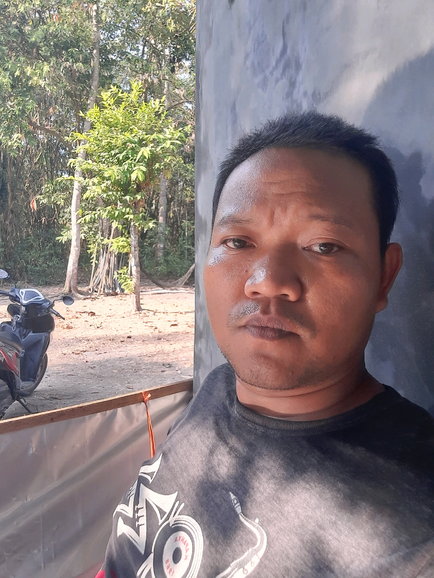 A man is taking a selfie outdoors in front of a building and a wooded area. He has brown skin, dark hair, and is wearing a dark t-shirt. The background shows a motorcycle, trees, and a gray wall. The scene is likely in Lugu, Indonesia.