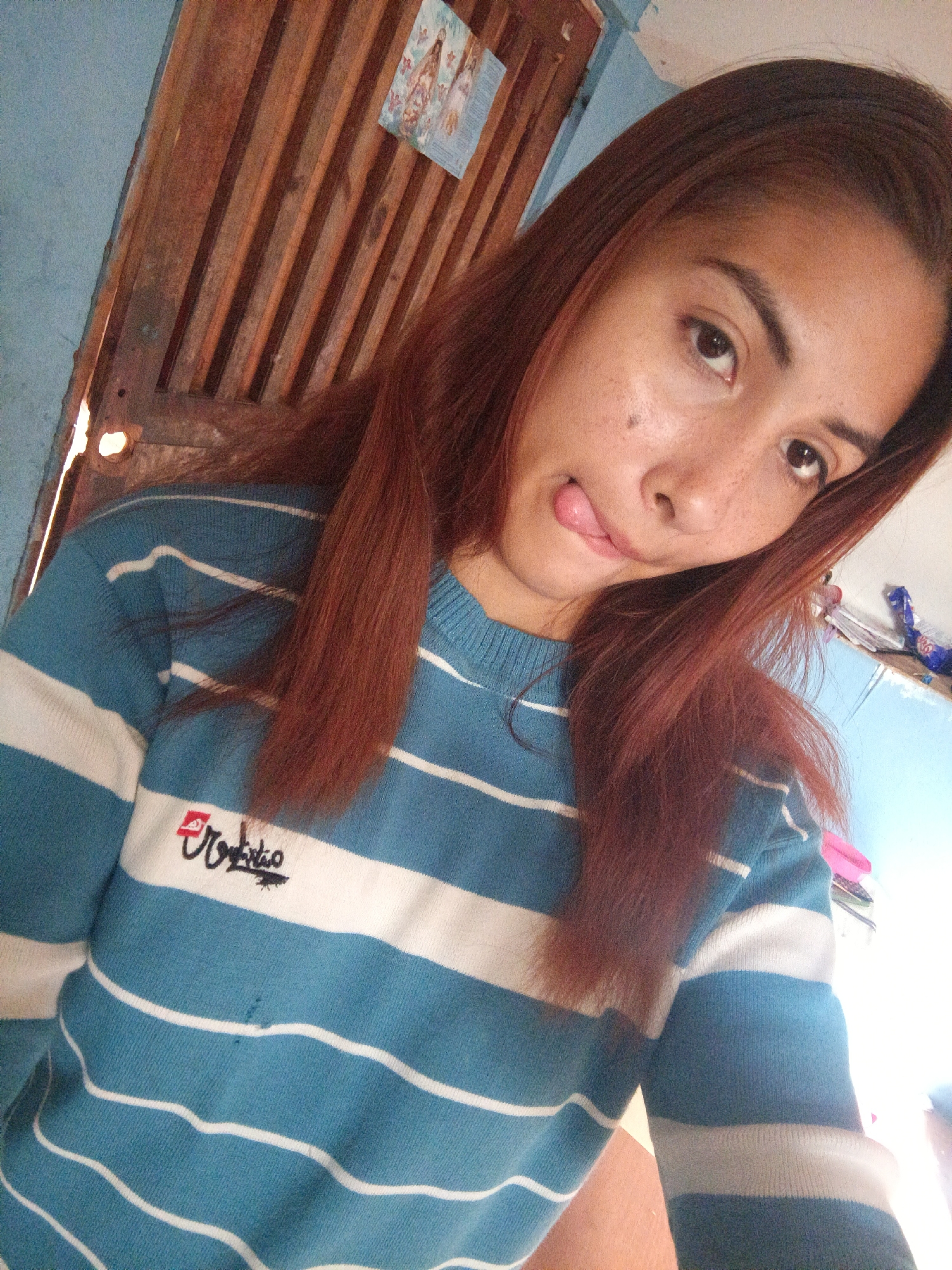 A woman with reddish-brown hair is posing for a selfie. She is wearing a blue and white striped sweater with embroidery on the chest. She is sticking her tongue out. The background includes a wooden door or wall, and blue walls, one of which has a small picture or calendar hanging on it. It is unclear where in Juan Griego, Venezuela this photo was taken.