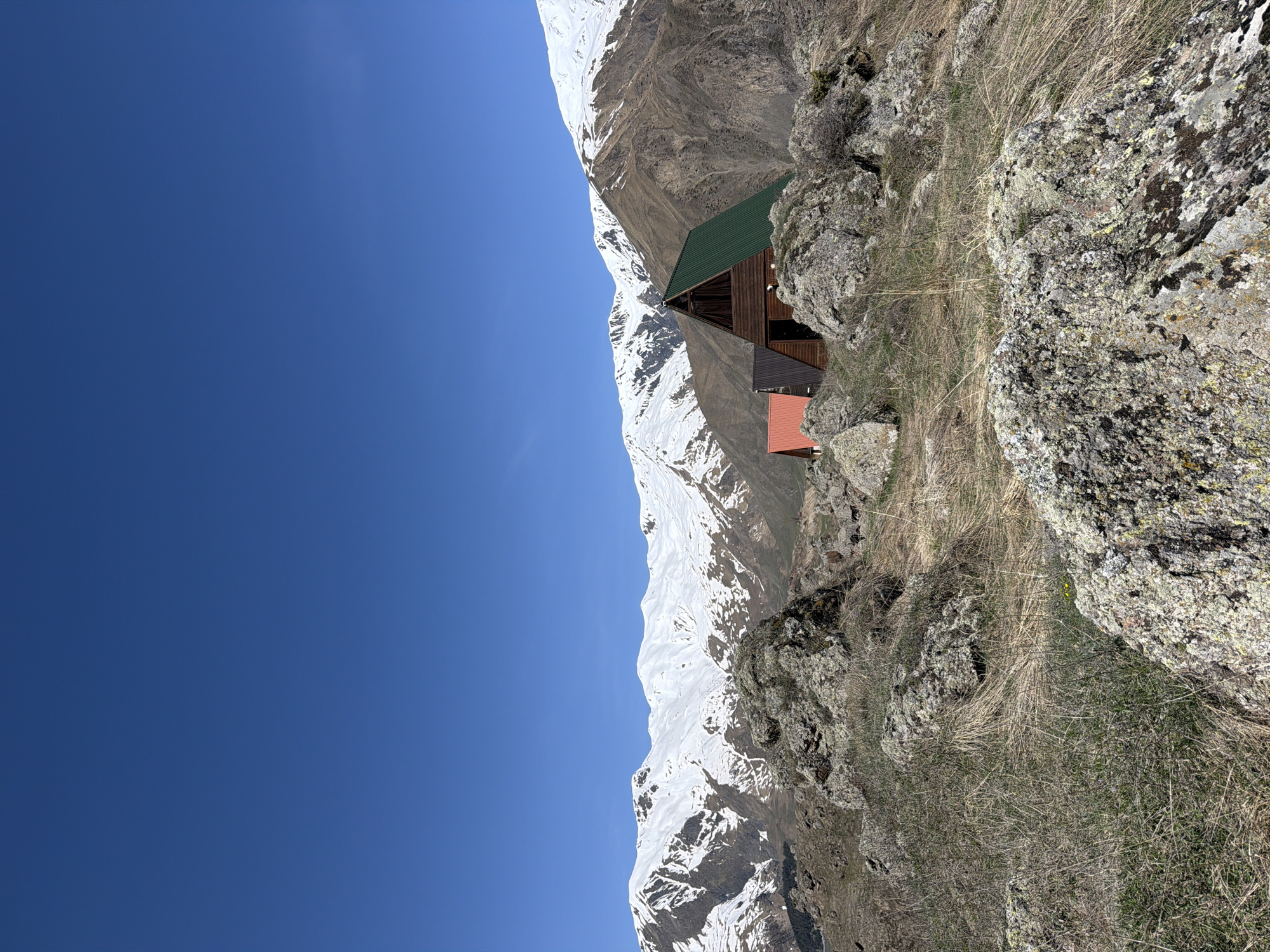 The image shows a mountain landscape with a building complex nestled on a rocky slope. Patches of snow cover the higher elevations of the mountains, set against a clear blue sky. The building complex consists of at least three structures with green, brown, and red roofs respectively. The buildings are situated on a rocky terrain with sparse vegetation. The location is likely near the city of Goristsikhe, Georgia.