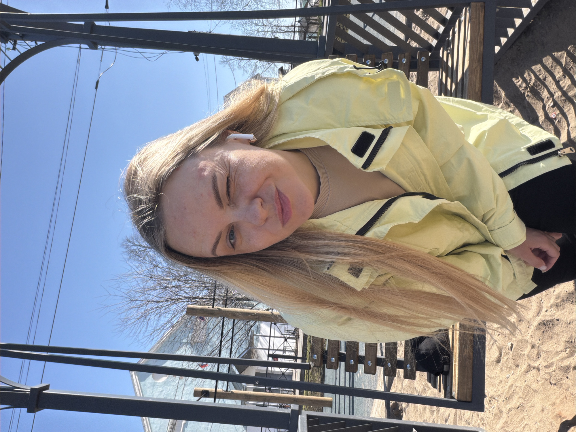 A woman with blond hair is outdoors in Votkinsk, Russia, likely at a playground or park. She is wearing a pale yellow jacket and an in-ear headphone. The playground equipment consists of metal and wood structures. The ground around the equipment appears to be sand. The sky is blue. Bare trees are visible in the background, indicating it is likely early spring.