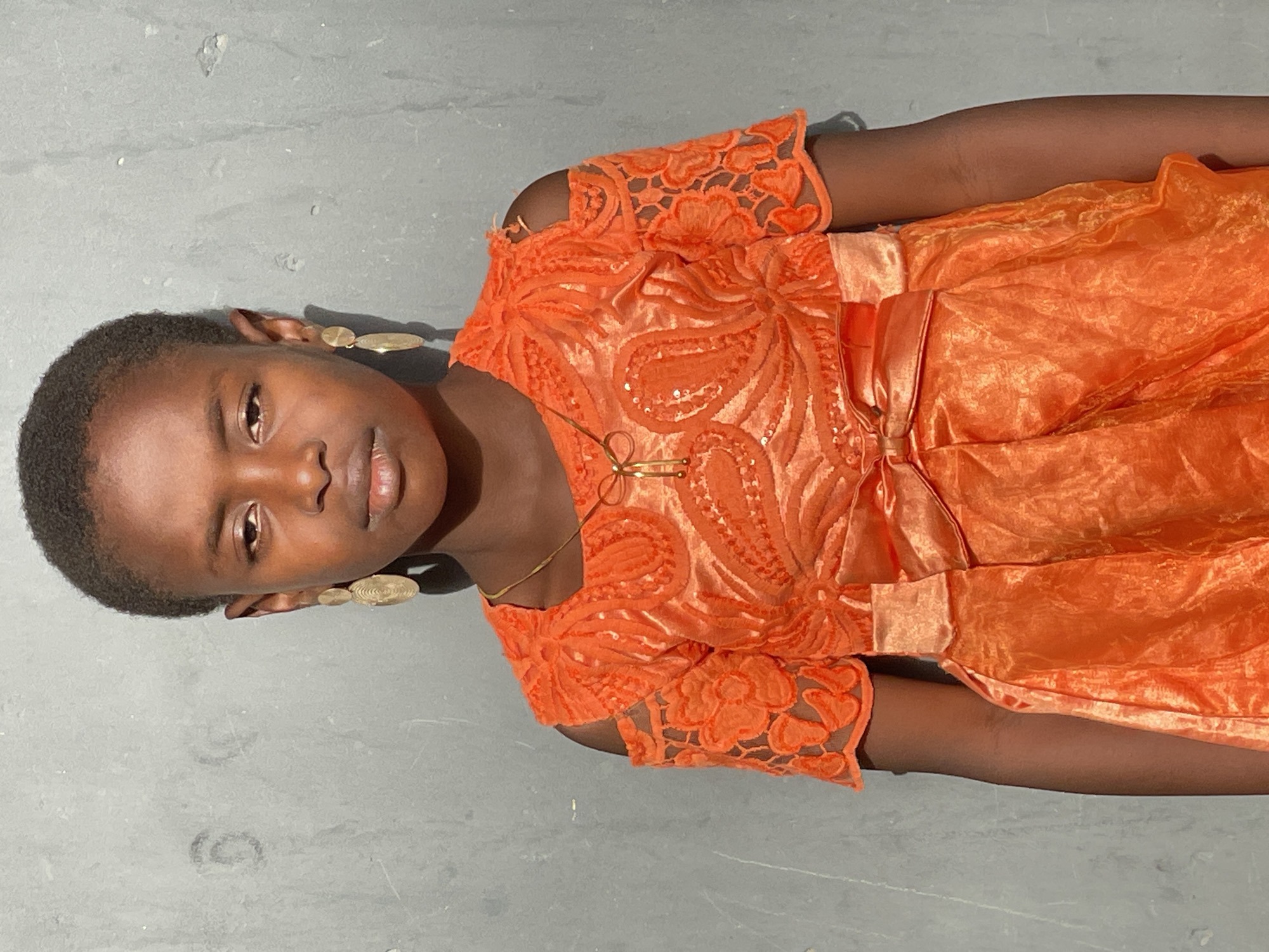 A girl with dark skin and short hair is wearing an orange dress with a lace bodice and a gold necklace. She also has gold earrings. She is posing indoors in front of a gray background. There is no indication of the city of Badagry, Nigeria.