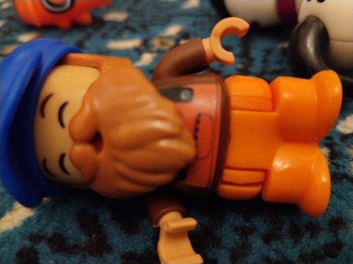 A small toy figure with a blue hat and beard lies on a textured surface. The figure has a tan-colored head with curved lines resembling eyes and a small hole for a mouth. It wears a brown shirt and bright orange pants and boots. One hand is raised in a curved "C" shape. Other toy figures are partially visible in the background.