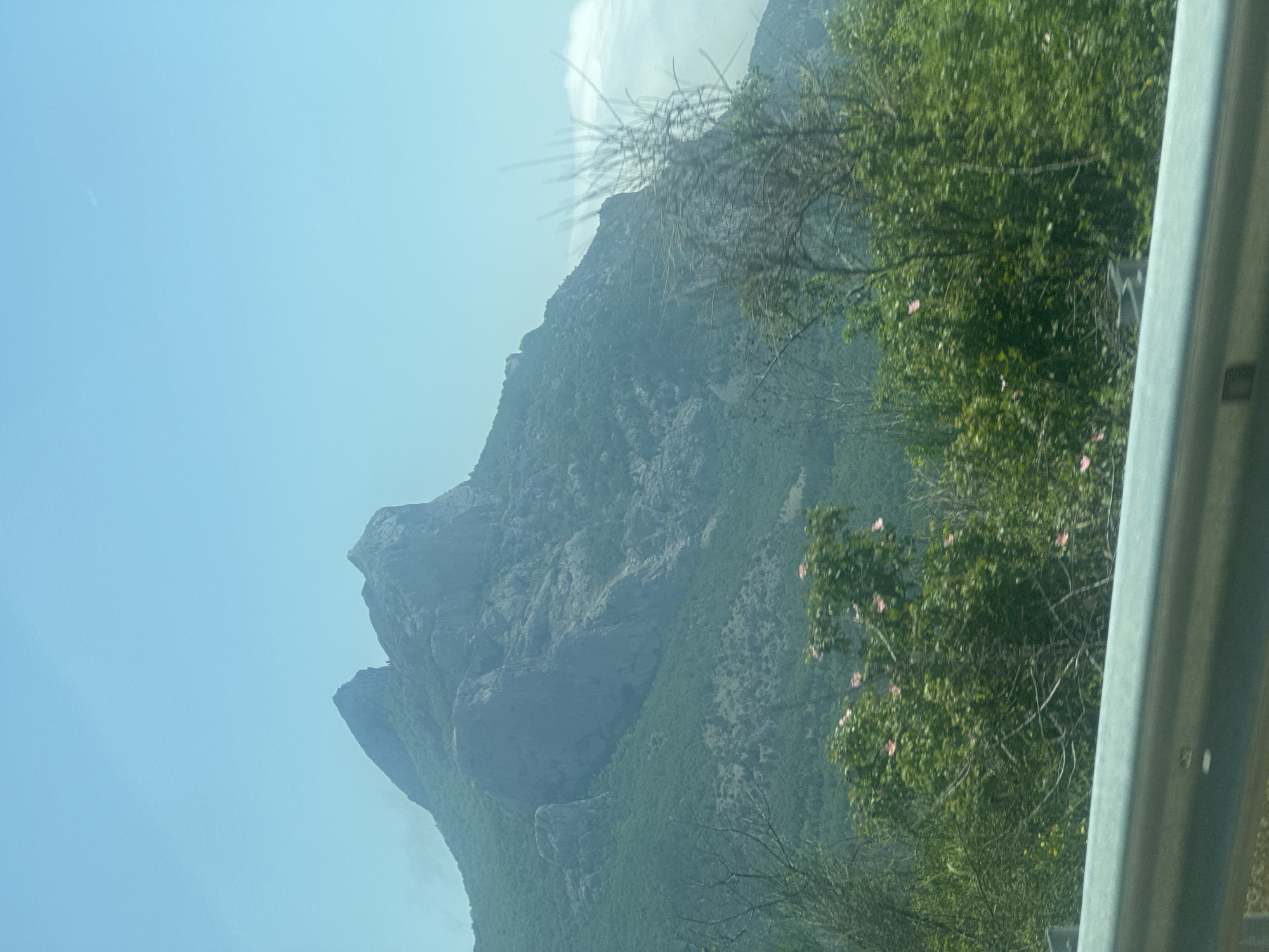 A landscape view showing a tall mountain range with rocky formations, partially shrouded in mist or low clouds. The sky is a light blue, and a tree with green foliage is visible on the right side of the frame. A gray guardrail is in the bottom right corner. Location: Unknown, Unknown.