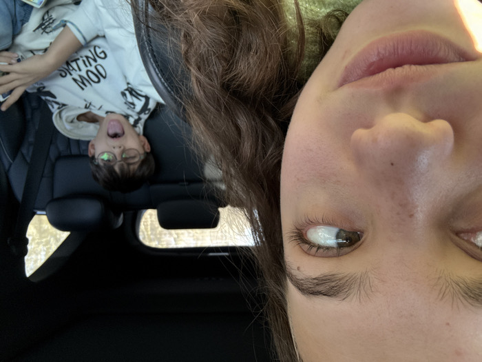 The scene depicts the interior of a vehicle in Banichan, Bulgaria, during daytime. In the left foreground, the right side of a female's face and upper torso are visible. She has medium-brown, wavy hair and is wearing a light green, textured garment. Her visible eye is directed towards the right side of the frame. In the right background, a male child is seated in a dark-colored rear passenger seat, secured by a shoulder seatbelt. He has short dark hair and wears round spectacles. He is dressed in a light-colored hooded sweatshirt with inverted dark text reading "SKATING MOD". His mouth is open, tongue extended, and his right eye is closed. His left hand is positioned as if holding a small electronic device. Blurred foliage and natural light are discernible through the vehicle's rear windows.